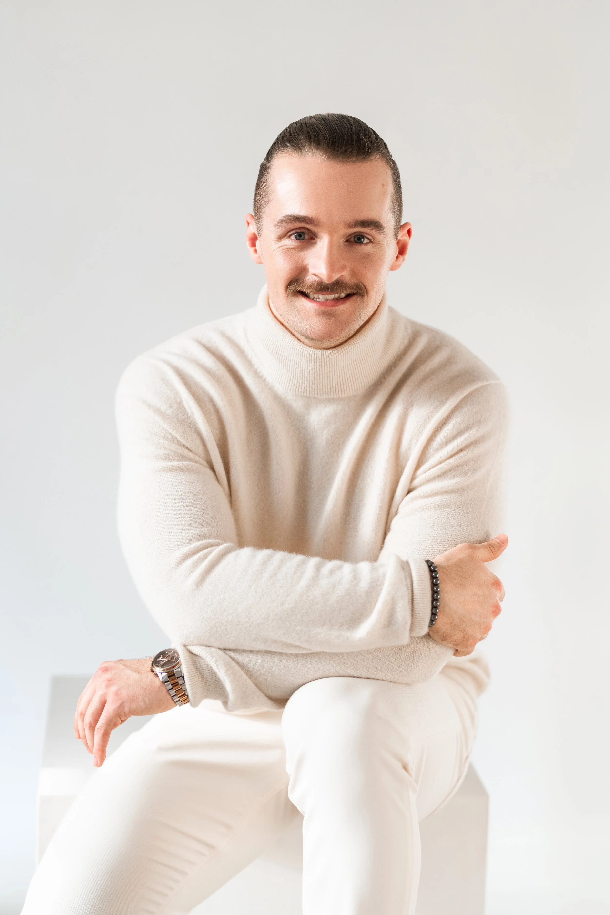 A professional corporate headshot of a man with dark hair, a mustache, and light skin, wearing a cream-colored turtleneck sweater and white pants, is smiling and sitting on a white stool against a plain light background.