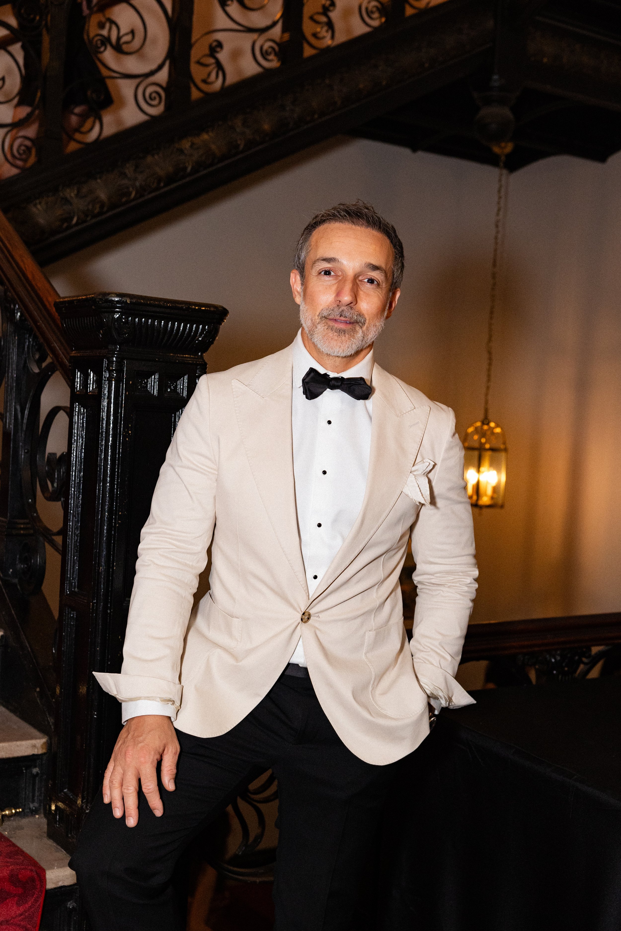 A corporate event professional photo of a man dressed in a tuxedo with a cream-colored blazer, white shirt, and black bowtie, posing indoors near a staircase with a black ornate railing, illuminated by warm lighting.