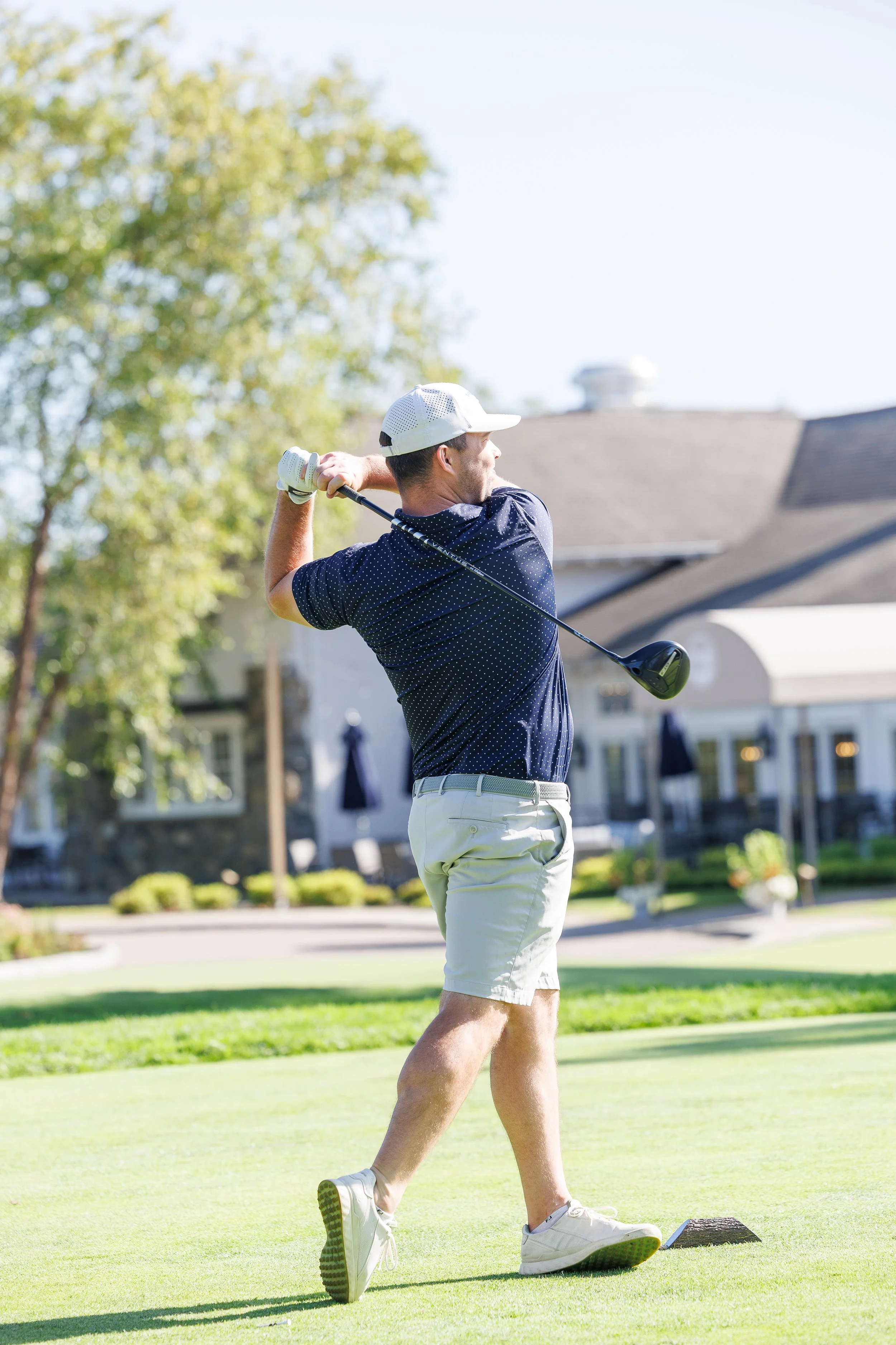 A corporate event professional photo of a man playing golf on a sunny day, swinging a golf club on a golf course with houses and trees in the background.