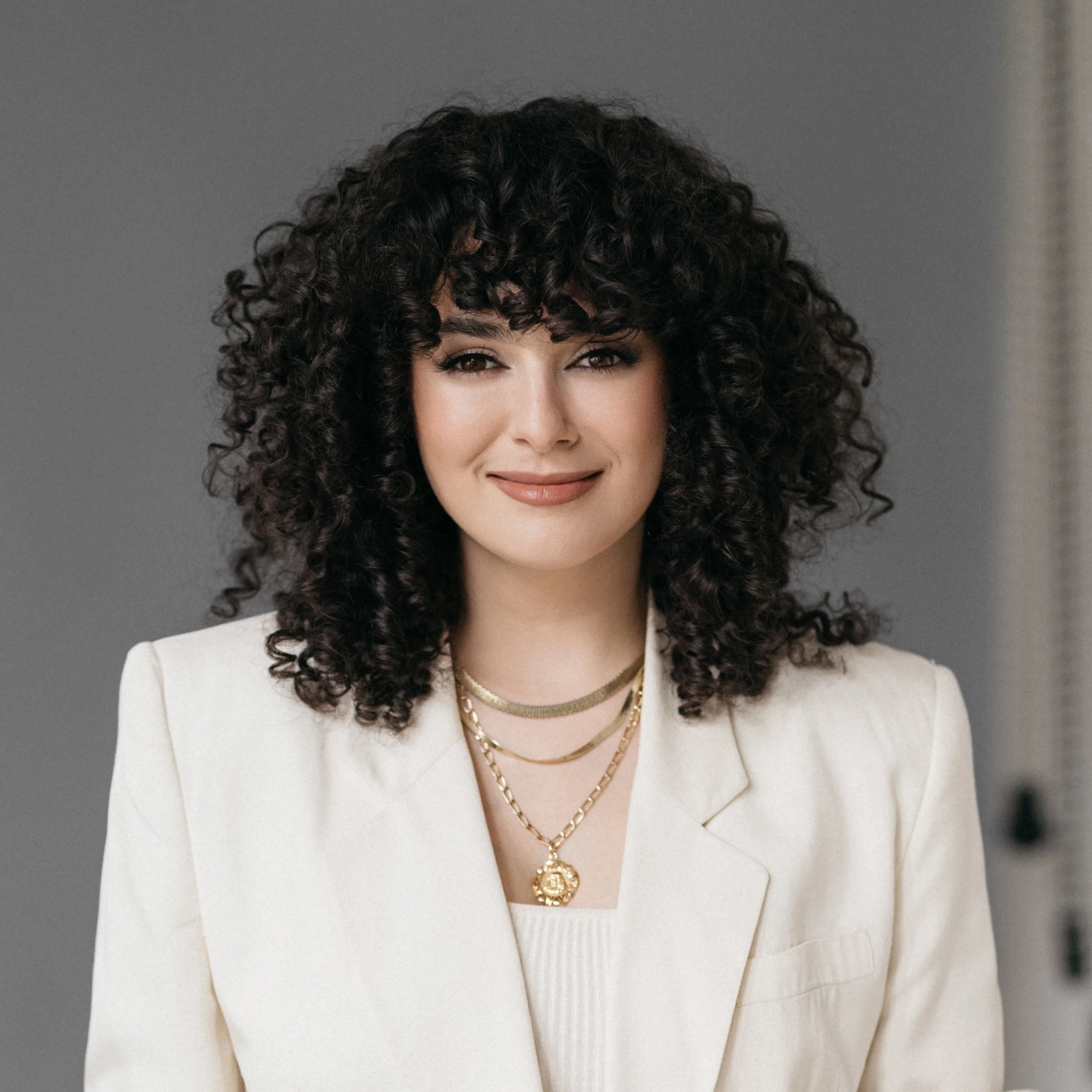 A professional corporate headshot of a close-up of a woman with curly brown hair wearing a white blazer and layered gold necklaces featuring a medallion pendant.