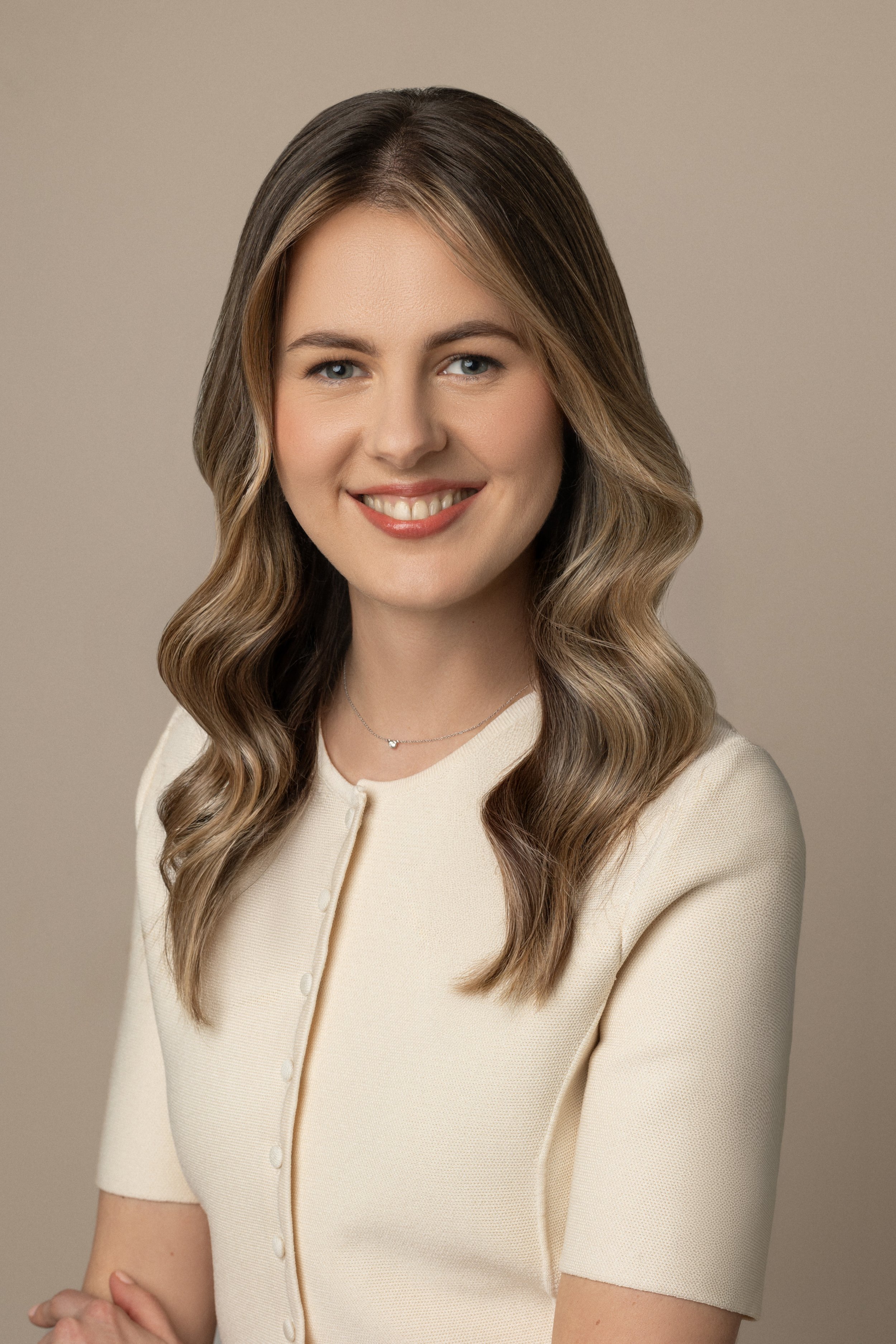 A professional corporate headshot of a woman with long wavy hair, wearing a cream-colored top and smiling at the camera against a neutral background.