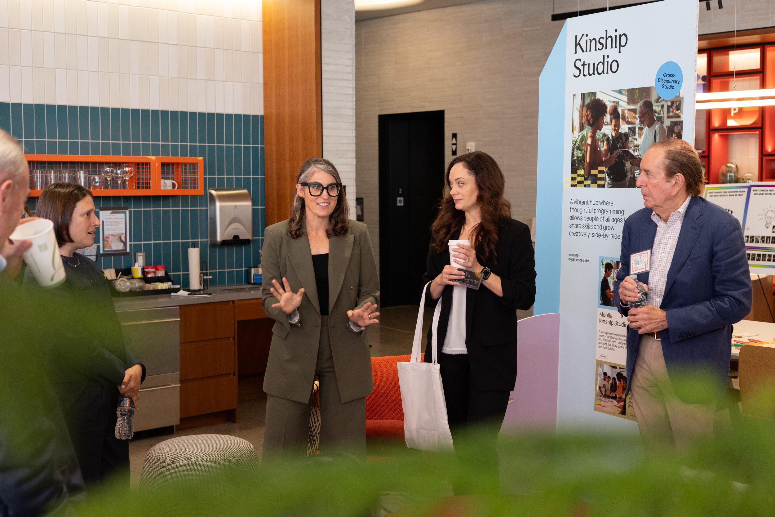A corporate event professional photo of People gathered at a conference or event near a large sign that reads 'Kinship Studio', with a subtitle about a cross-disciplinary studio for sharing skills and growth.
