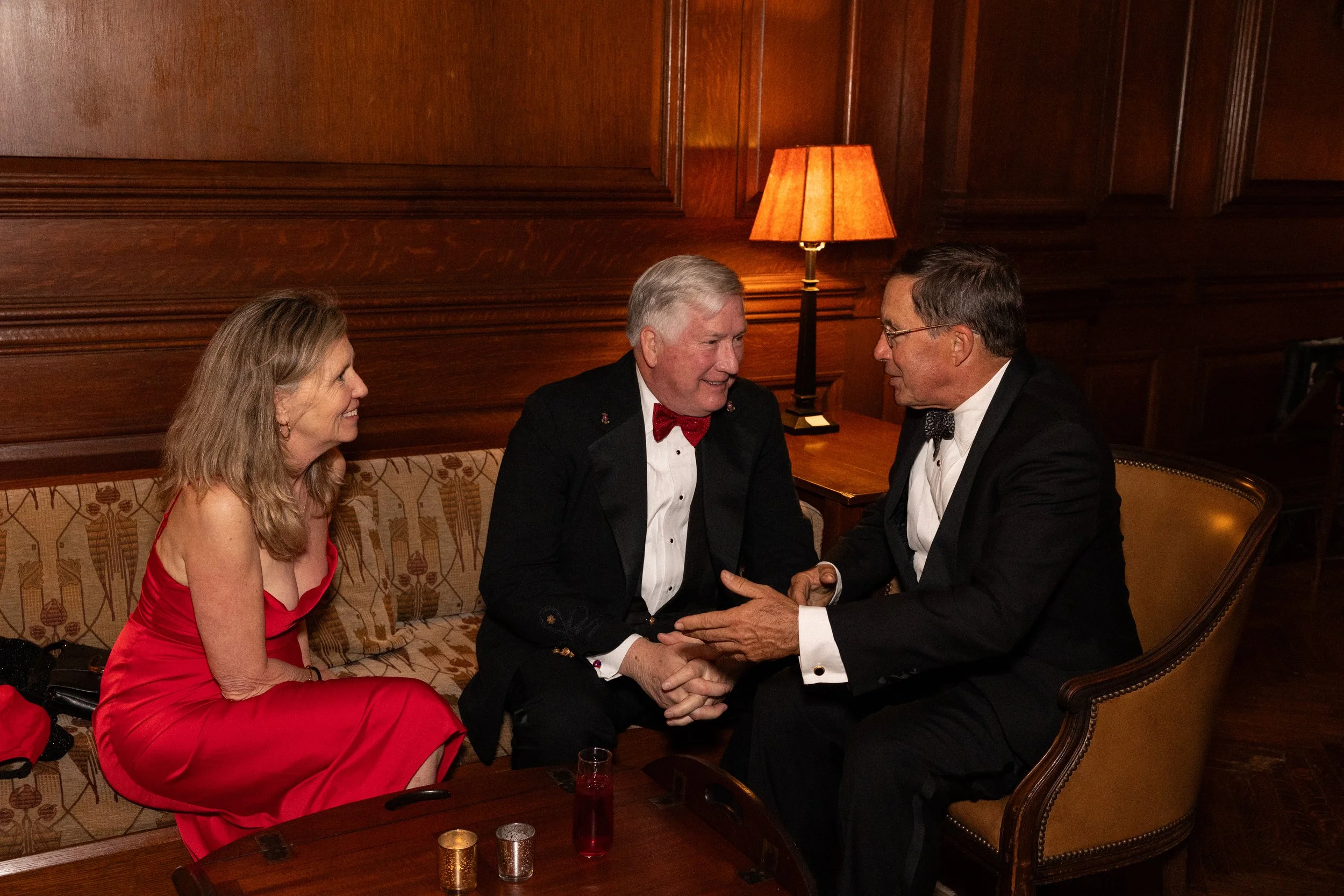 A corporate photo of three people, two men and a woman, sitting and conversing in a warmly lit wood-paneled room. One man and the woman are smiling, while the other man looks on. The woman wears a red dress, and both men wear tuxedos with bow ties.