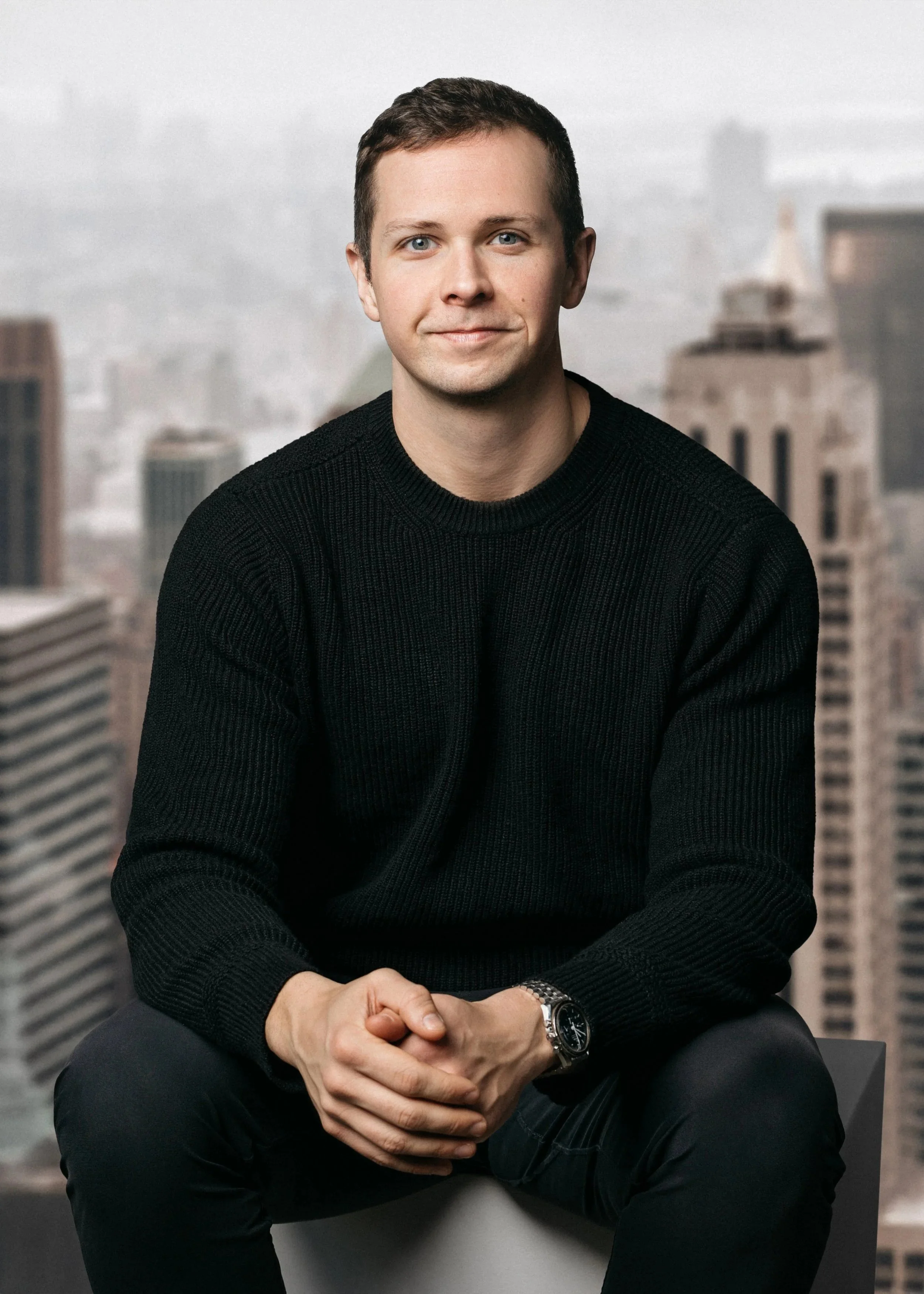 A professional corporate headshot of a young man with short brown hair and blue eyes, wearing a black sweater, sitting in front of a blurred cityscape background.