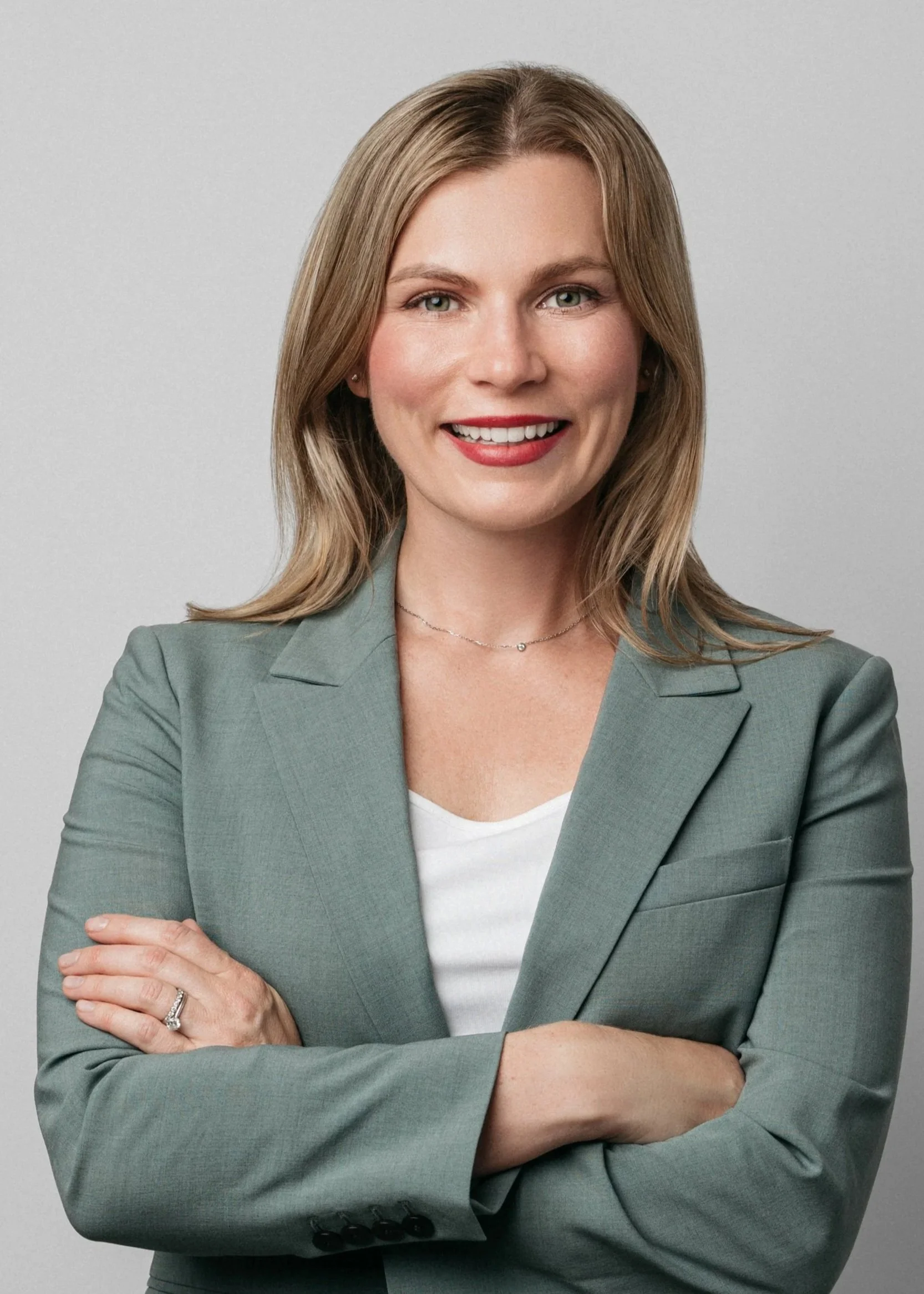 A professional corporate headshot of a woman with shoulder-length blonde hair, green eyes, and a friendly smile, wearing a light gray blazer over a white top, with arms crossed, standing in front of a light gray background.