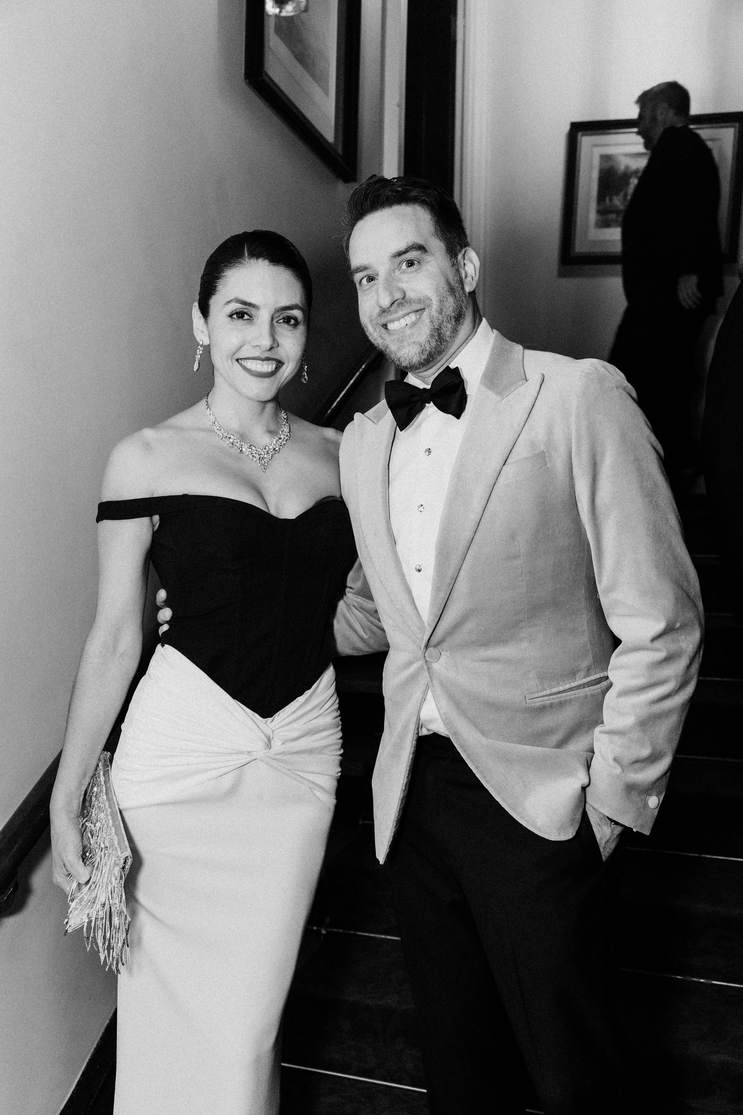 A black-and-white photo of a smiling woman in an off-shoulder black top and a woman in a white dress with a jacket, both dressed for a formal event, standing close together inside a room with paintings on the wall. In the background, a man in a dark suit is walking away.