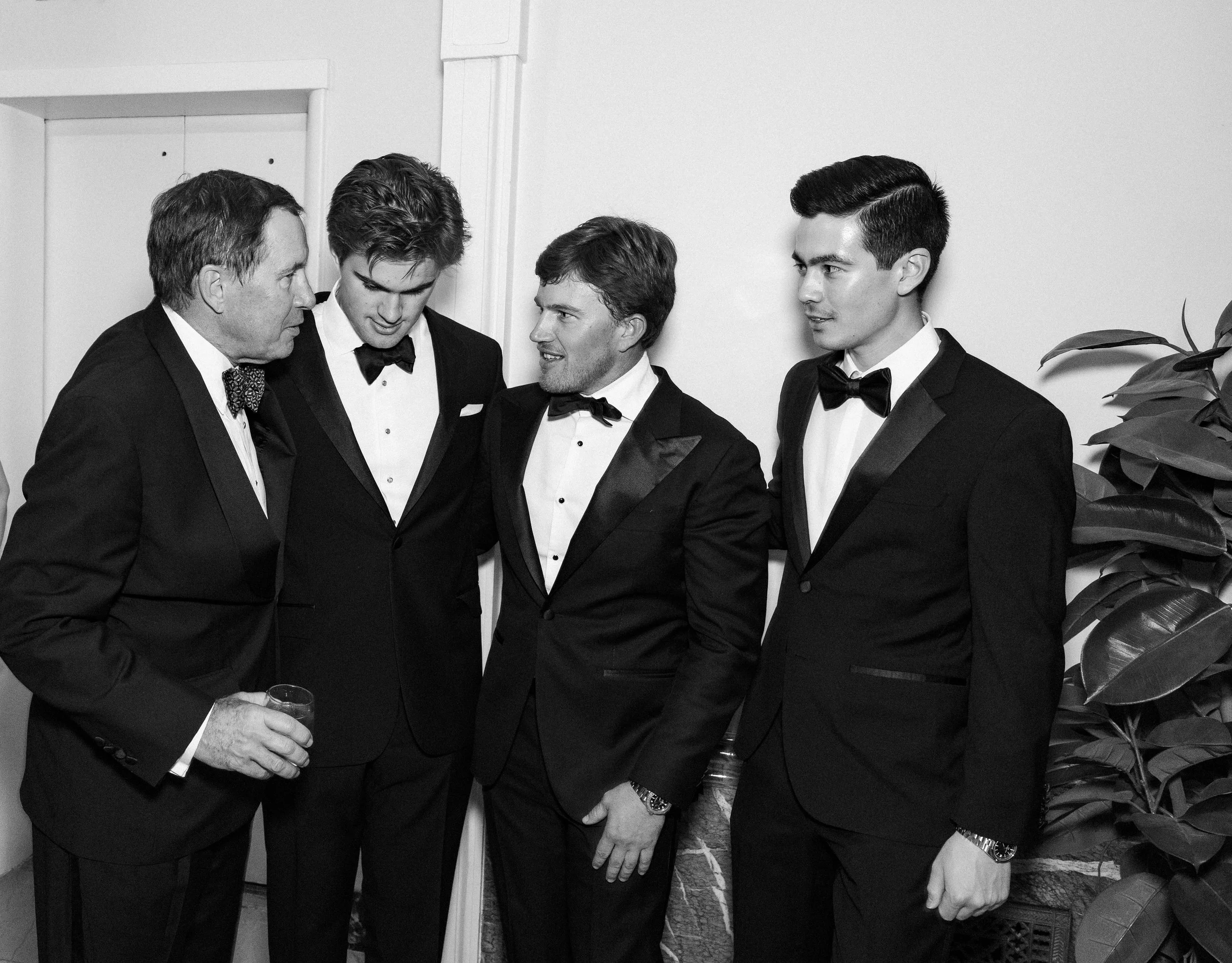 A corporate event professional photo of four men in tuxedos, two young and two older, standing together in conversation at a formal event, with one holding a drink in his hand.