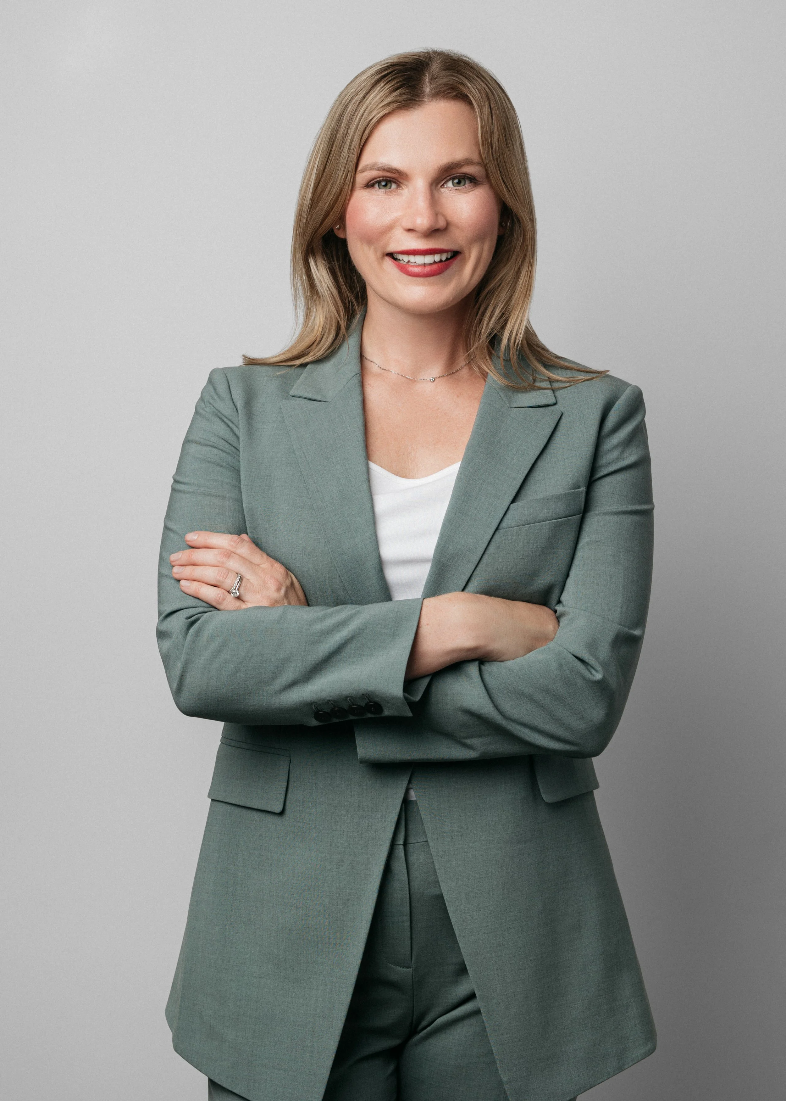A  corporate headshot of a woman with shoulder-length blonde hair, wearing a light green blazer and white top, smiling with crossed arms against a plain grey background.