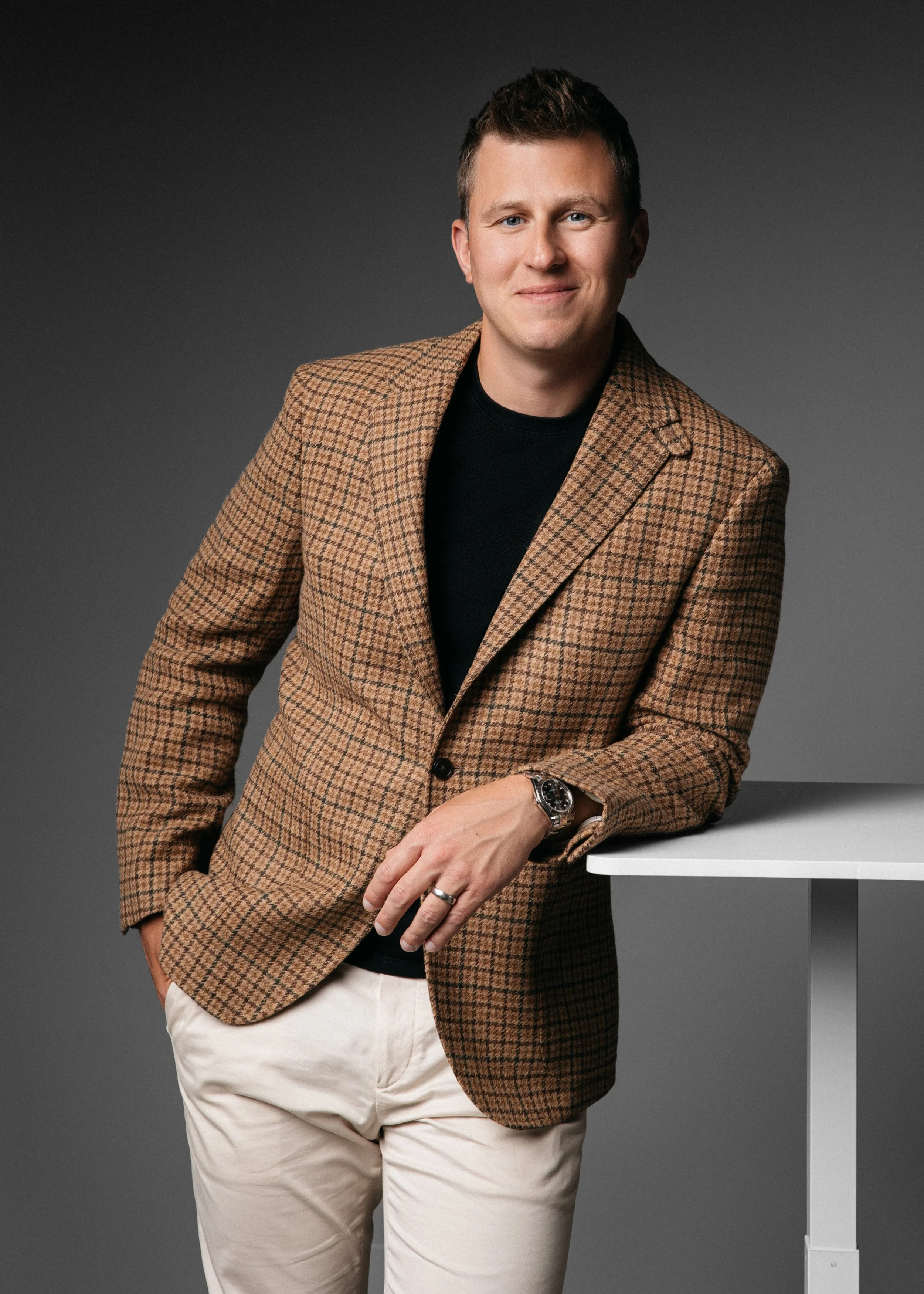 A headshot photo of a man wearing a brown plaid blazer and white pants, leaning on a white table against a plain gray background.