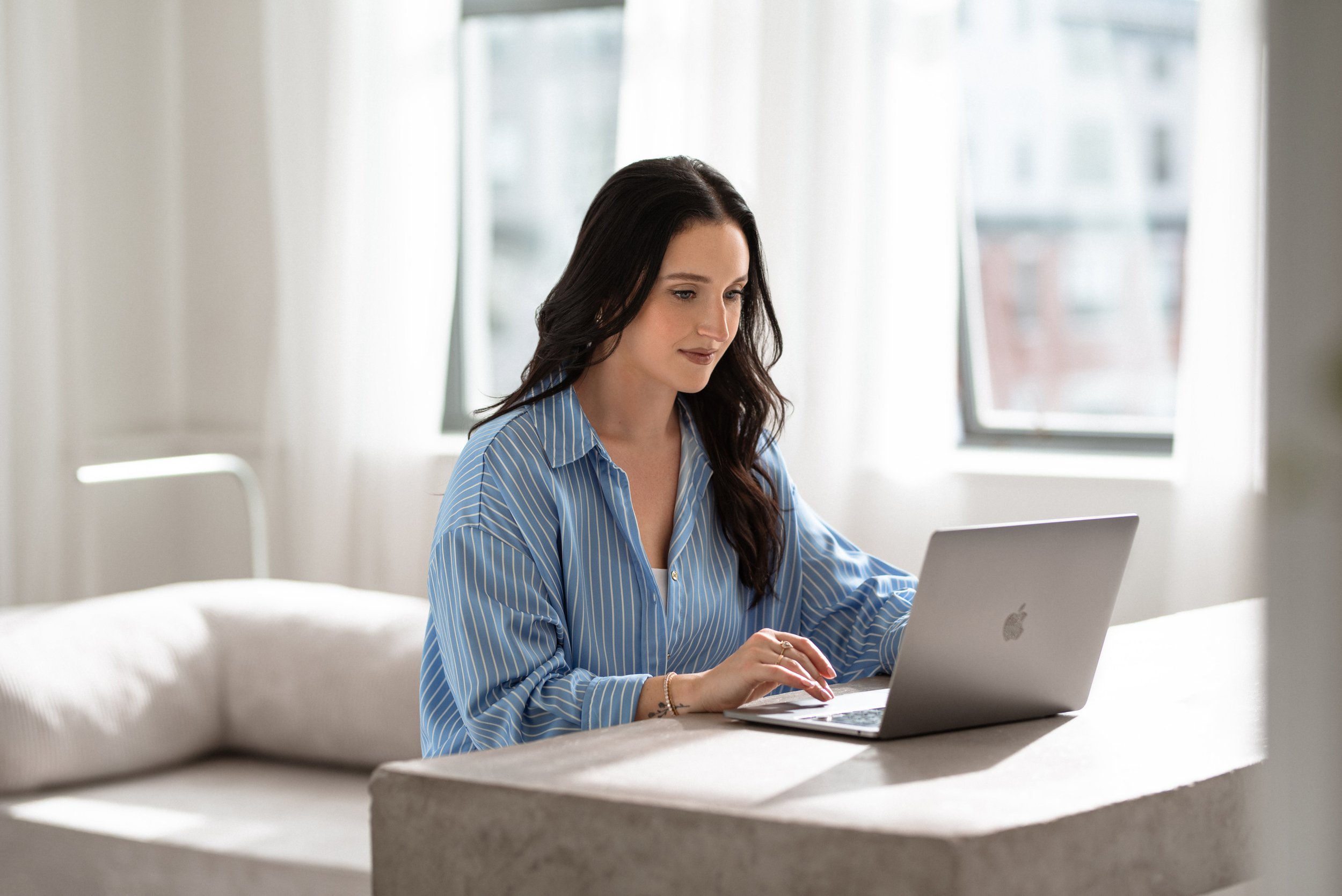 A professional corporate headshot of a woman with dark hair wearing a blue striped shirt working on a laptop at a table in a bright room with large windows.