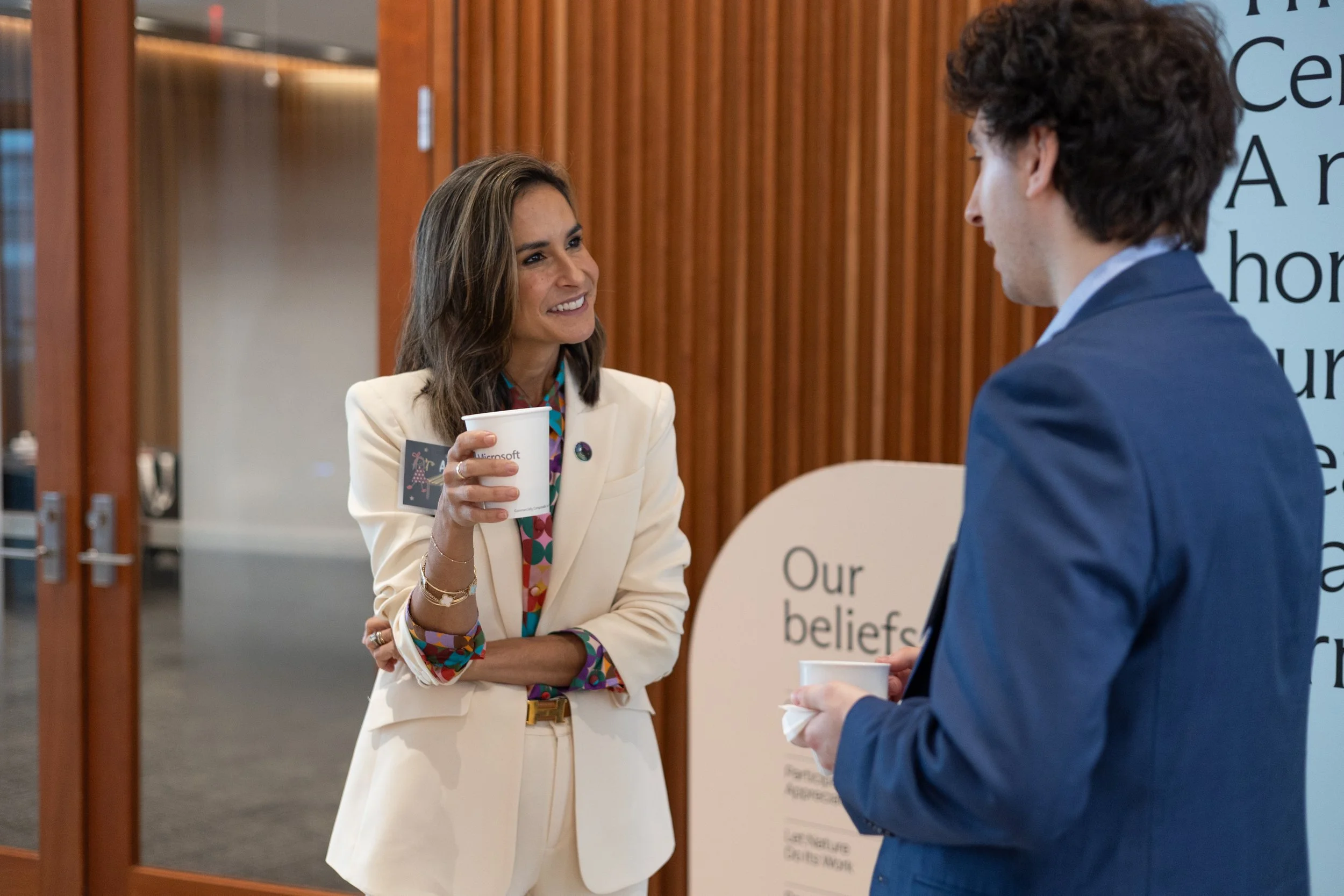 A corporate event photo of a woman and a man engaged in conversation in an indoor setting. The woman is smiling, holding a cup, and wearing a cream-colored blazer with colorful jewelry. The man is dressed in a blue suit.