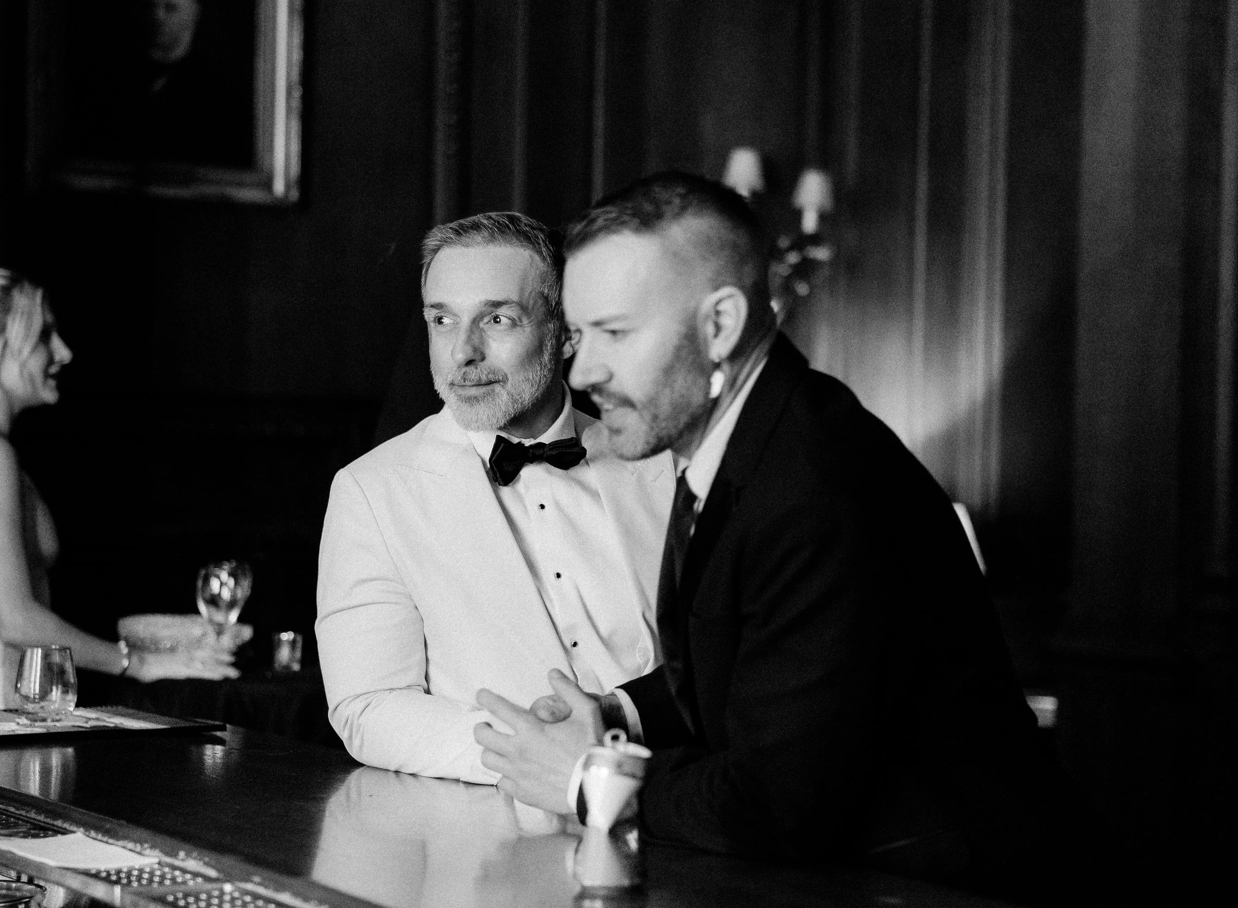 A corporate event professional photo of two men in formal attire, one in a tuxedo and the other in a white shirt with a bow tie, sitting at a table and engaging in conversation. The setting appears to be an elegant dinner or event.