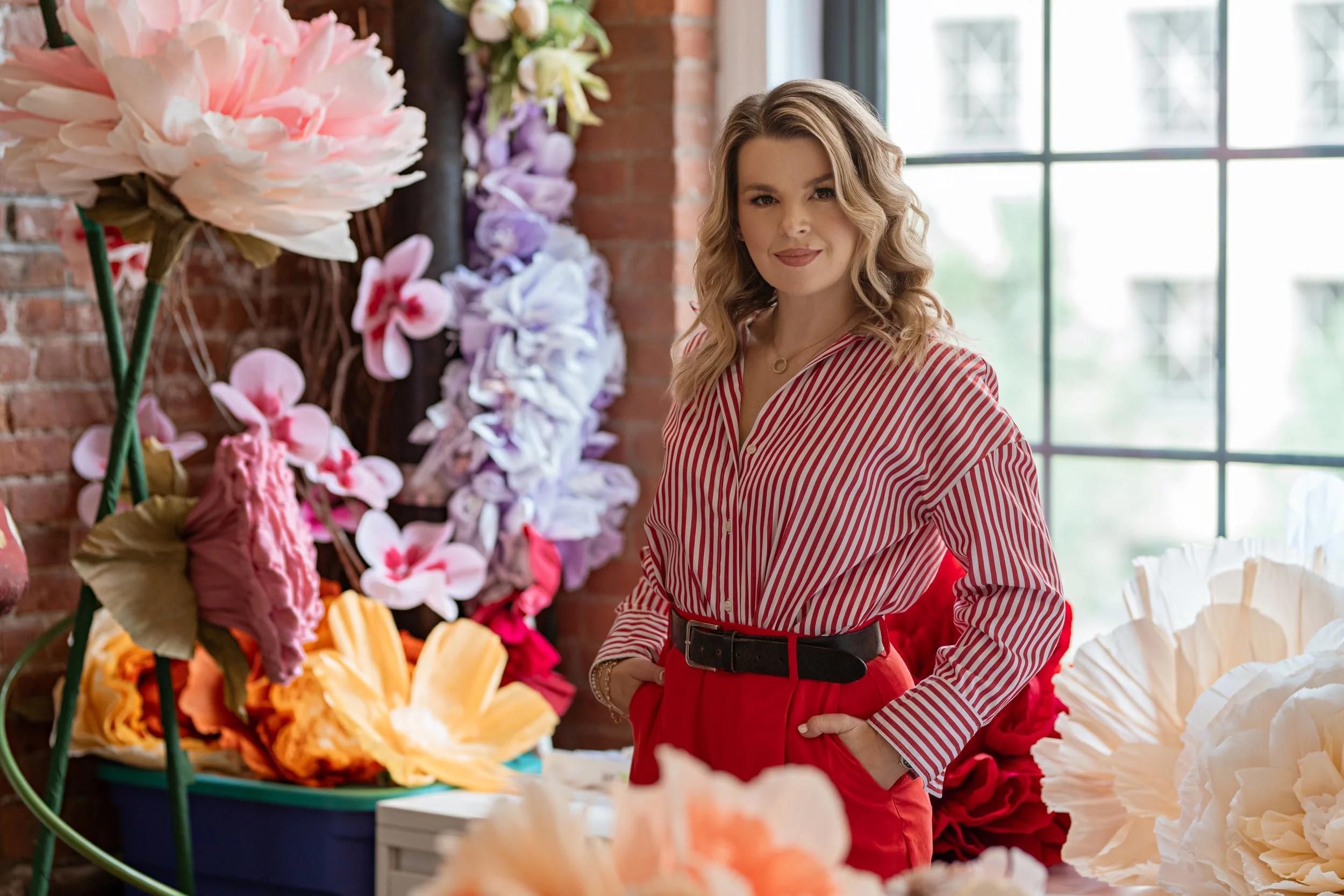 A professional corporate headshot of a woman with curly blonde hair wearing a red and white striped blouse and red pants standing in front of a table of large, colorful paper flowers in pink, orange, purple, and white.