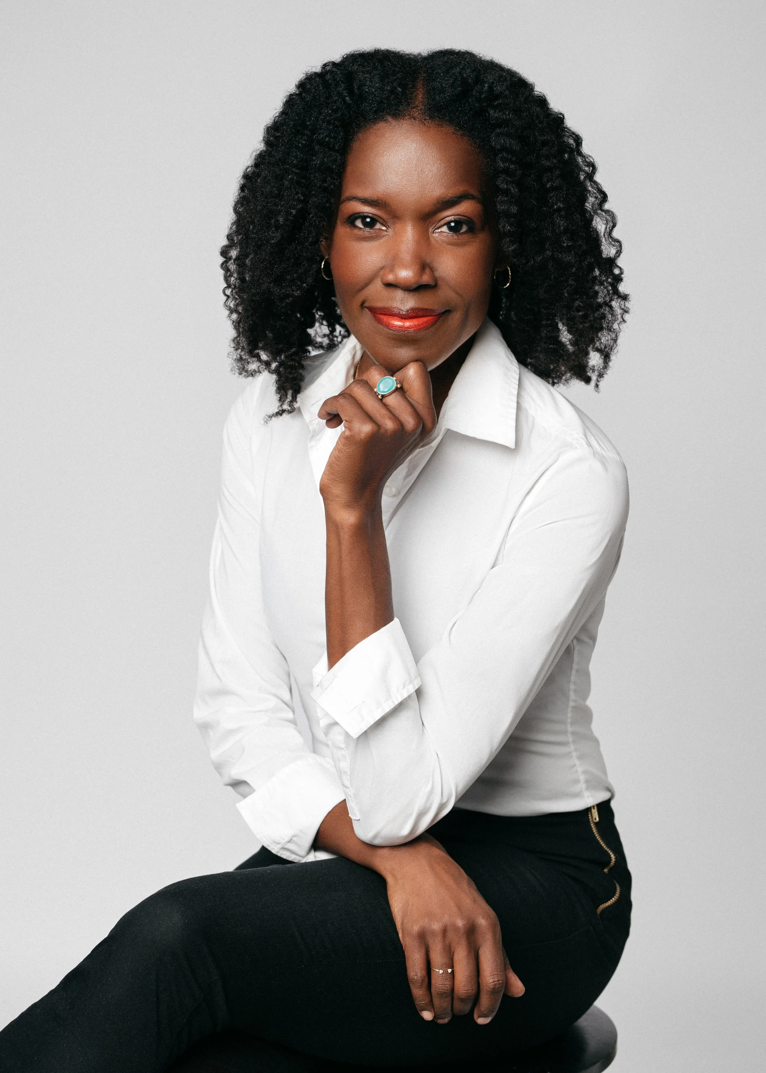A professional corporate headshot: Portrait of a woman with natural curly black hair, wearing a white shirt and black pants, sitting on a black stool against a plain light grey background, looking at the camera with a confident expression.