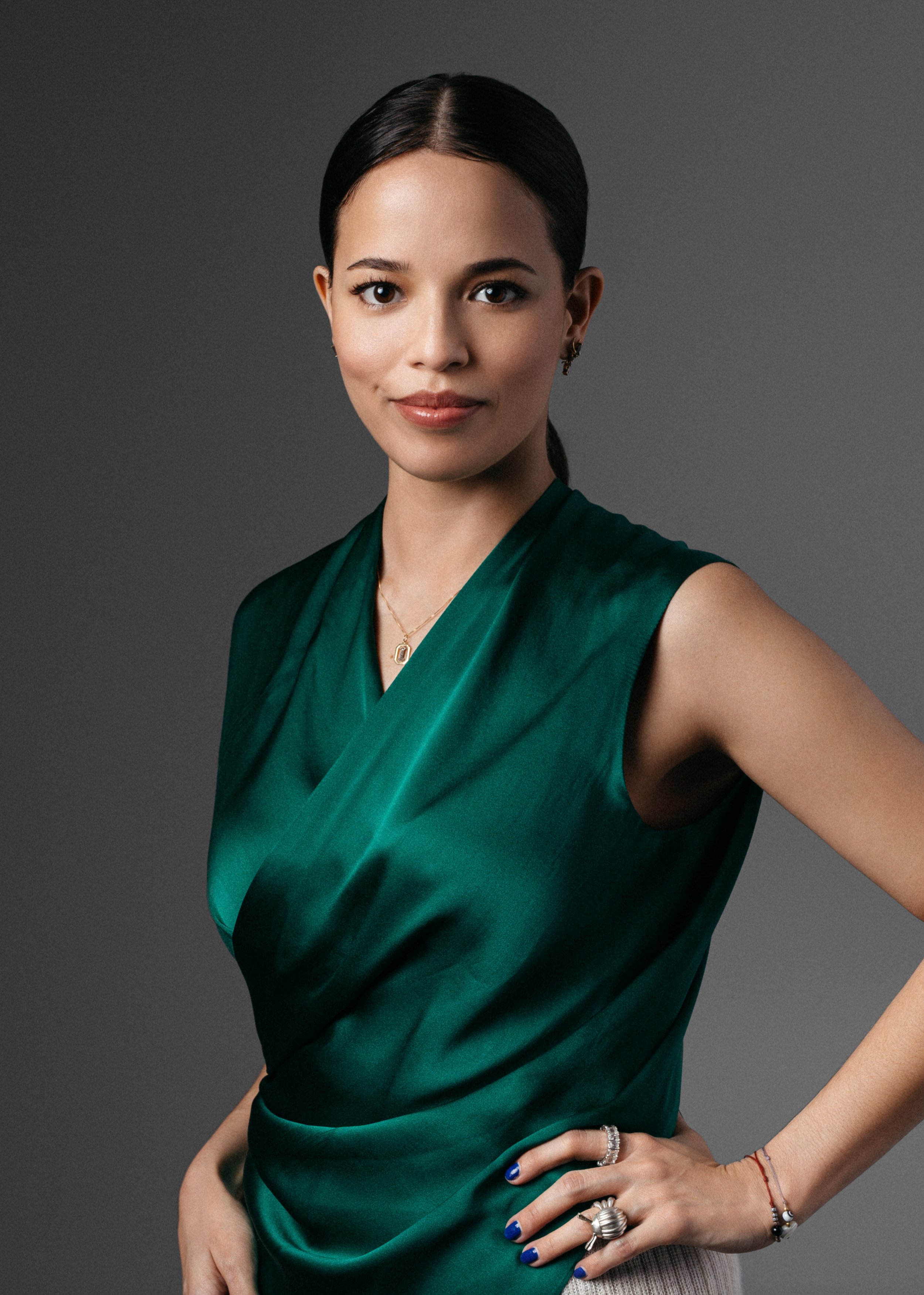 A  corporate headshot of a woman with dark hair pulled back, wearing a green satin sleeveless dress, holding her hip, wearing jewelry including rings, a necklace, bracelets, and earrings, against a gray background.