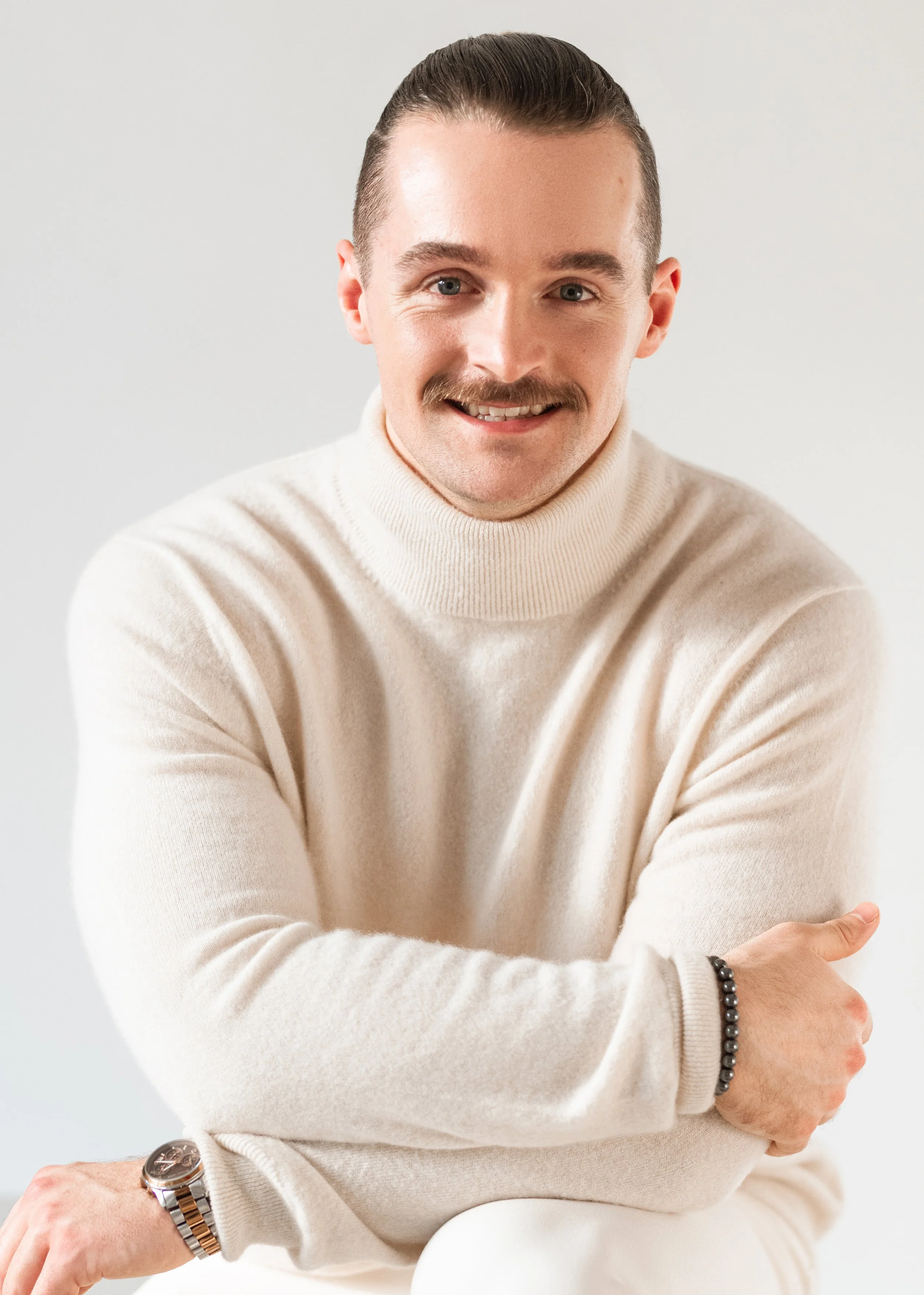A professional corporate headshot of a smiling man with slicked-back hair and a mustache, wearing a cream-colored turtleneck sweater, is sitting with his arms crossed, wearing a watch and bracelet, against a plain light background.