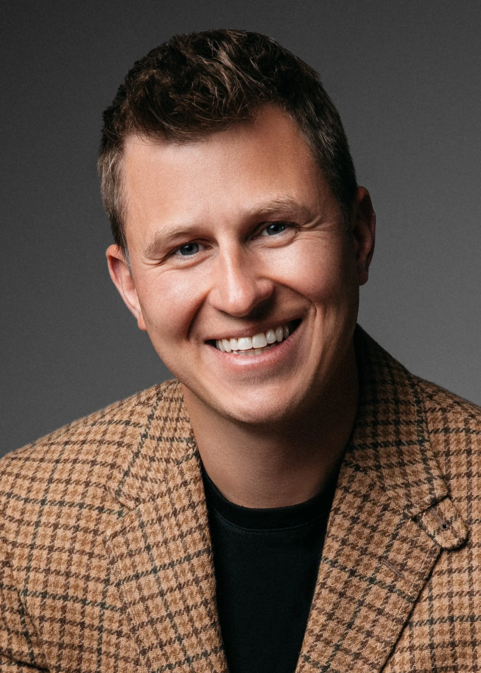 A professional corporate headshot of a Close-up of a smiling man with short dark hair, wearing a brown checkered jacket over a black shirt, against a dark gray background.