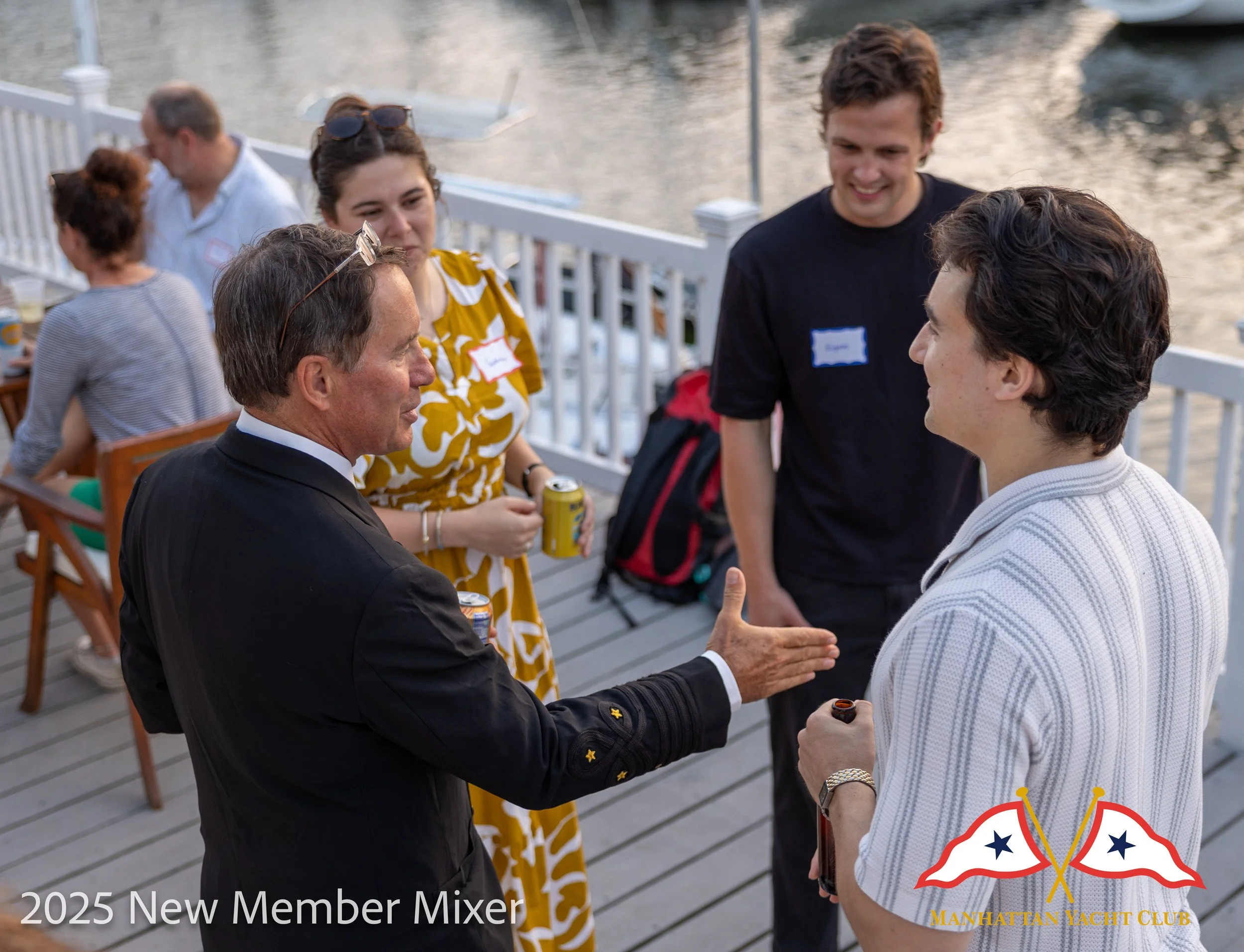 A corporate event professional photo of  a group of people at a social gathering on a deck near water, engaging in conversation. There are men and women, some wearing name tags, with drinks in hand. In the foreground, two men are shaking hands. 