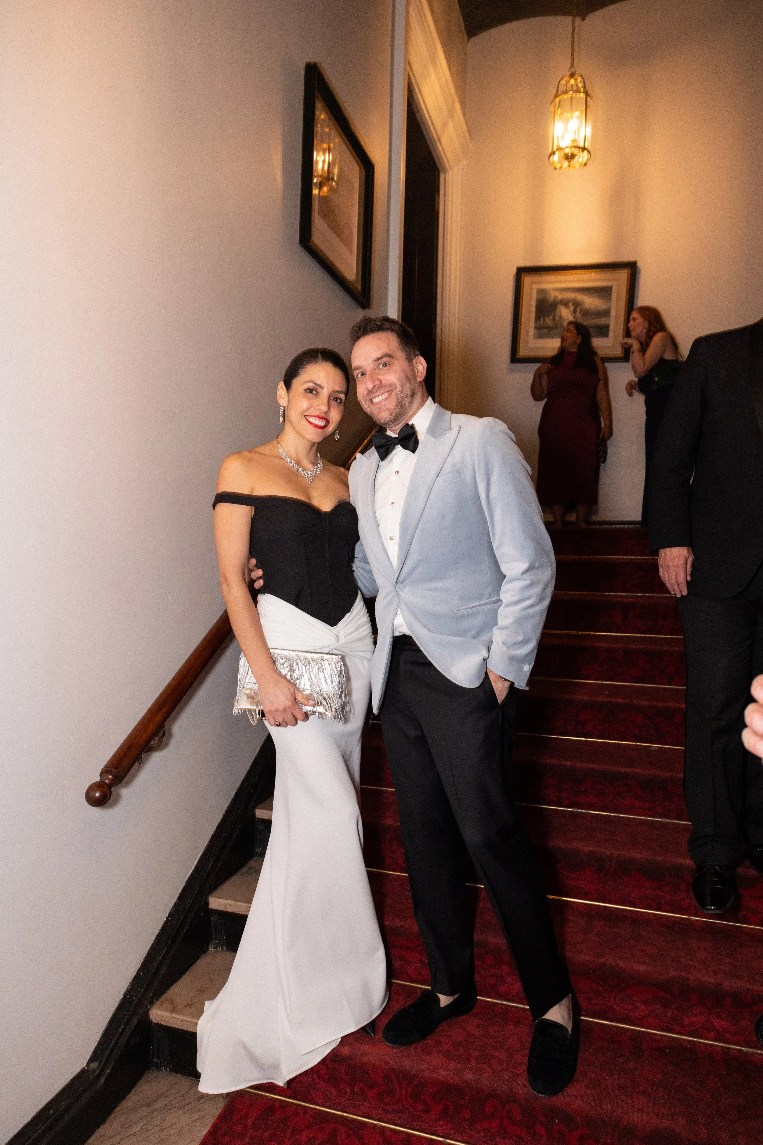 A corporate event professional photo of a woman and a man dressed in formal attire, standing on a staircase, smiling at the camera at an event or celebration.