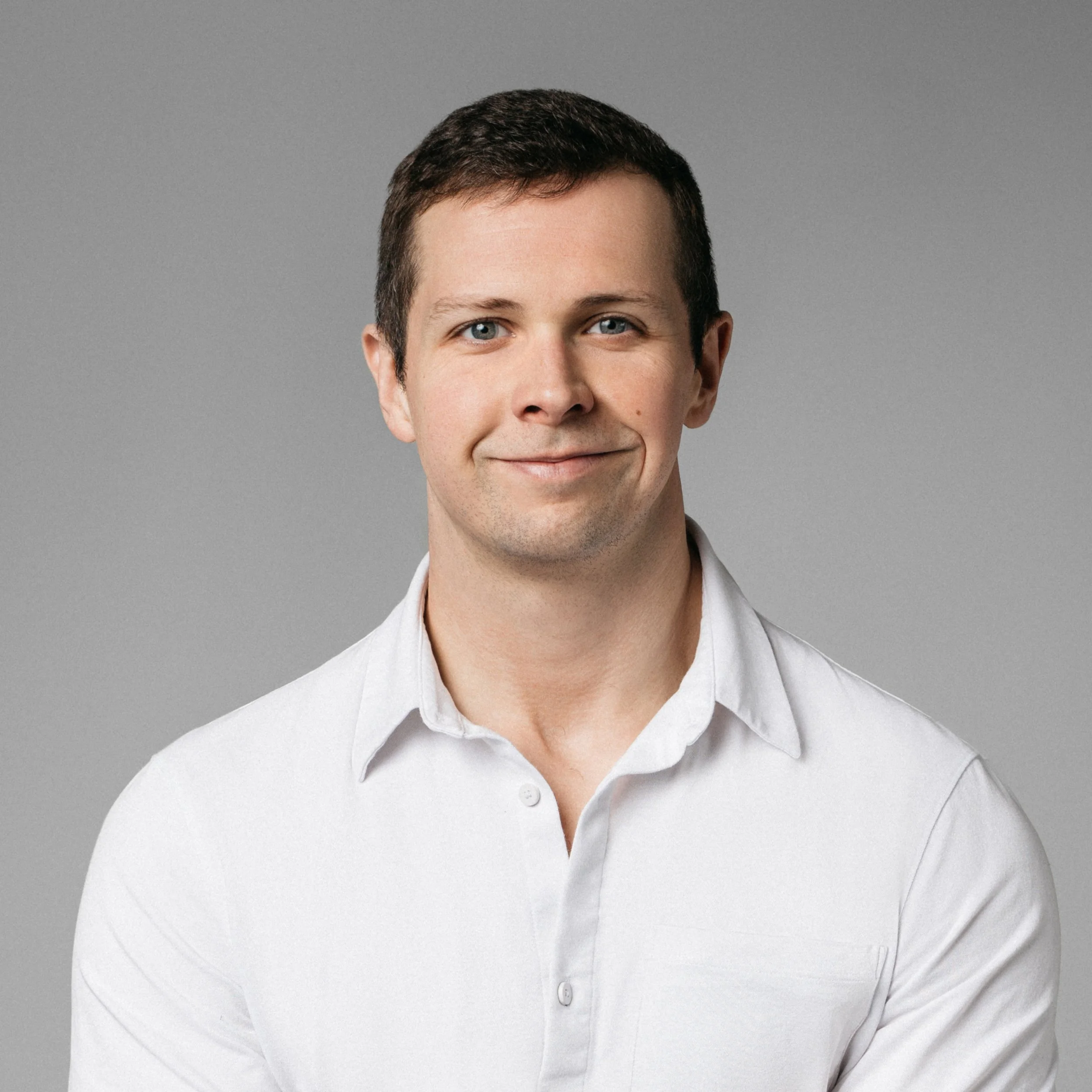 A professional corporate headshot of a close-up of a man with short brown hair and blue eyes, wearing a white button-up shirt, smiling subtly against a gray background.