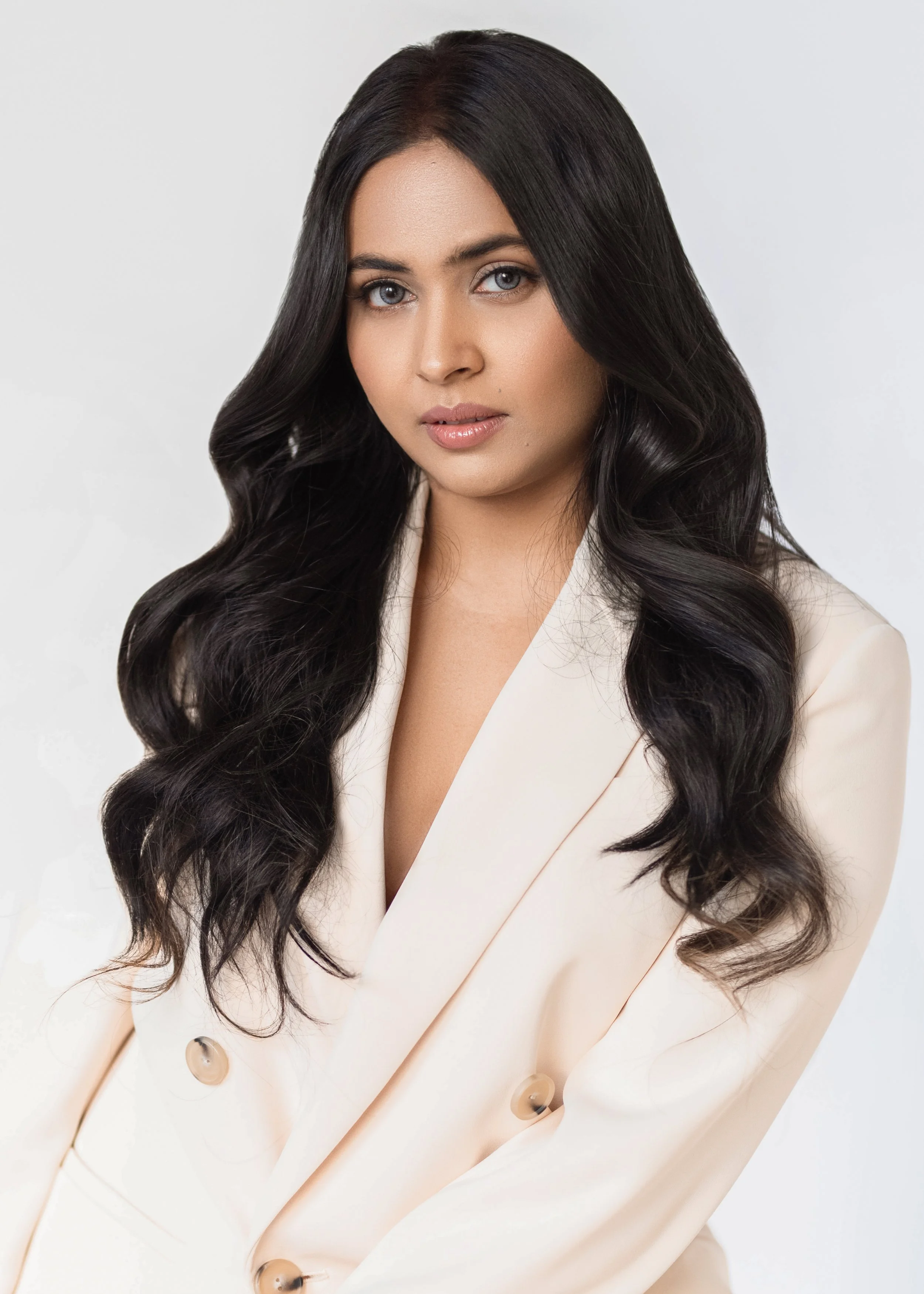 A professional corporate headshot of a woman with long, wavy black hair and light makeup wearing a light-colored blazer.