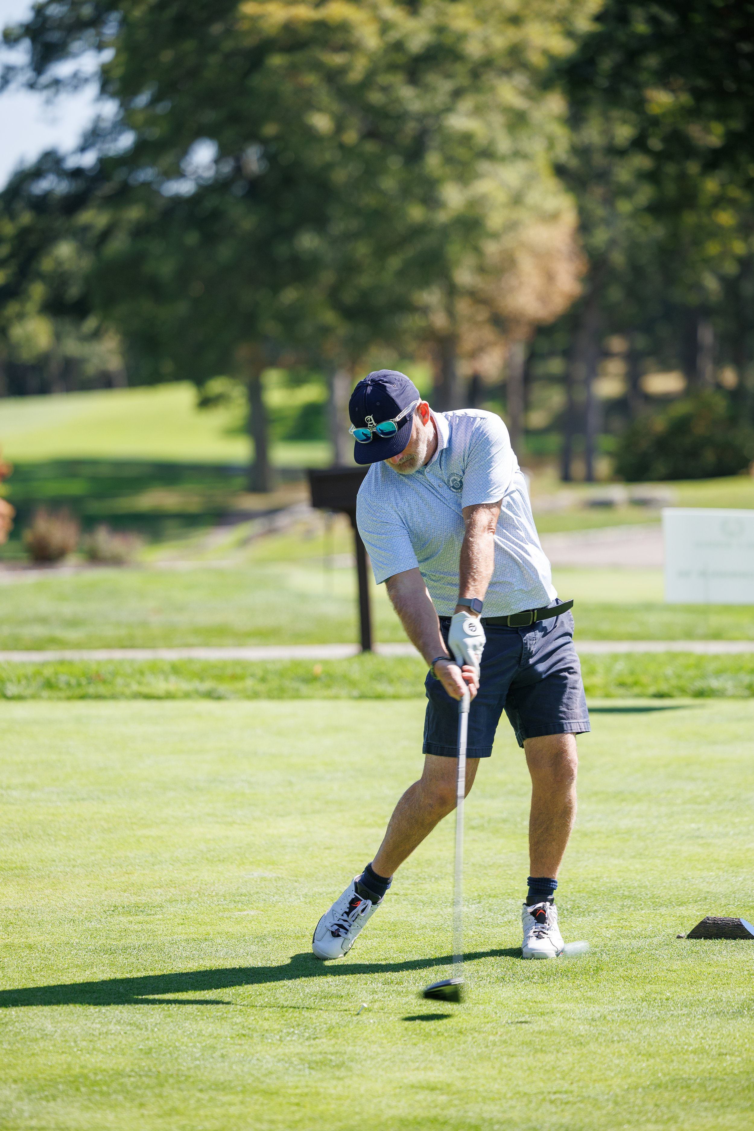 A corporate event professional photo of A man on a golf course wearing a white shirt, dark shorts, white golf shoes, a black cap with goggles, practicing putting with a golf club. The background shows trees and a sunny day.