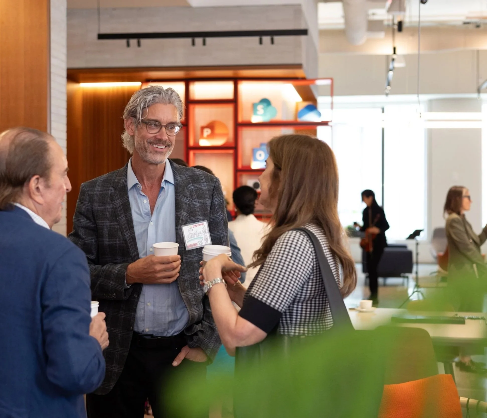 A corporate event photo of a group of mature professionals in business at a modern office or conference space, with one man talking and smiling, while women and other men listen. The background features colorful shelving with letters and other decor.