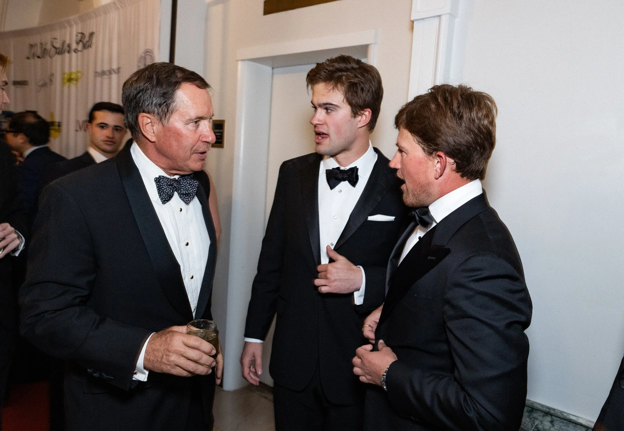 A corporate event professional photo of three men in tuxedos having a conversation at a formal event, one holding a drink.
