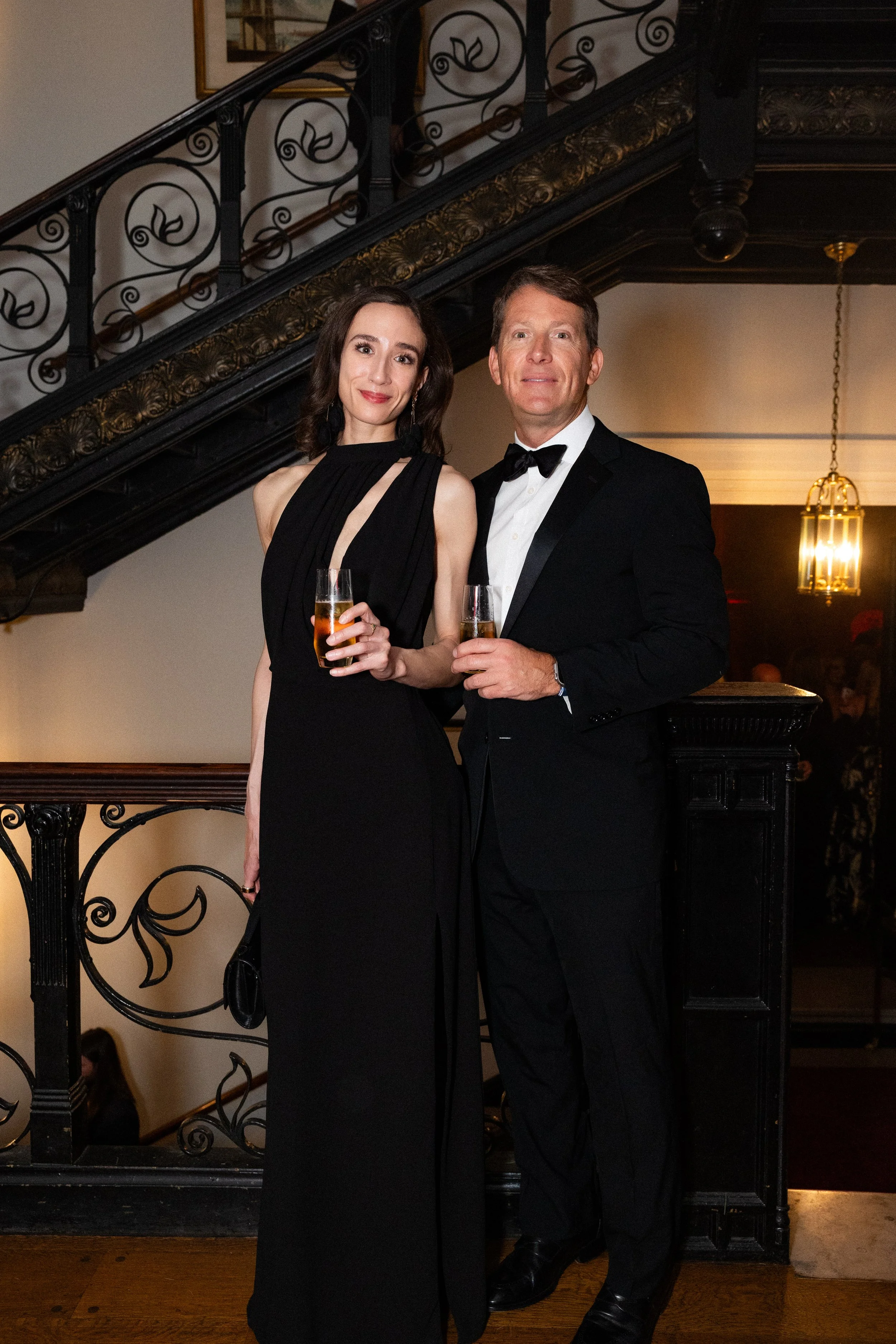 A corporate event professional photo of a man and woman dressed in formal attire, holding champagne glasses, standing indoors near a staircase with an ornate black and gold railing, and warm lighting in the background.