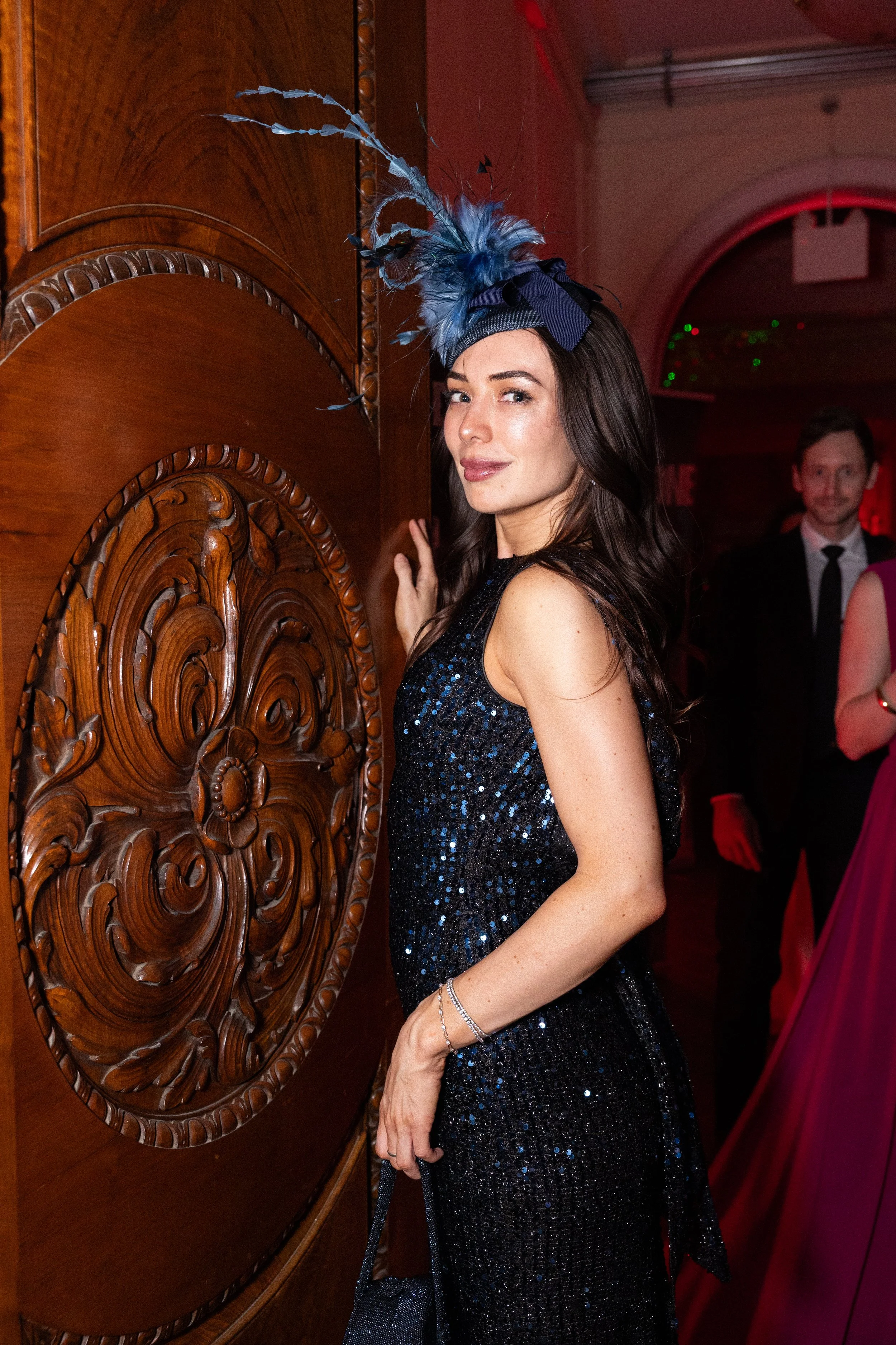 A professional event photo of a woman wearing a blue feathered fascinator and a sparkly black dress poses at a formal event, standing next to an ornate wooden door with intricate carvings, with other people in formal attire in the background.