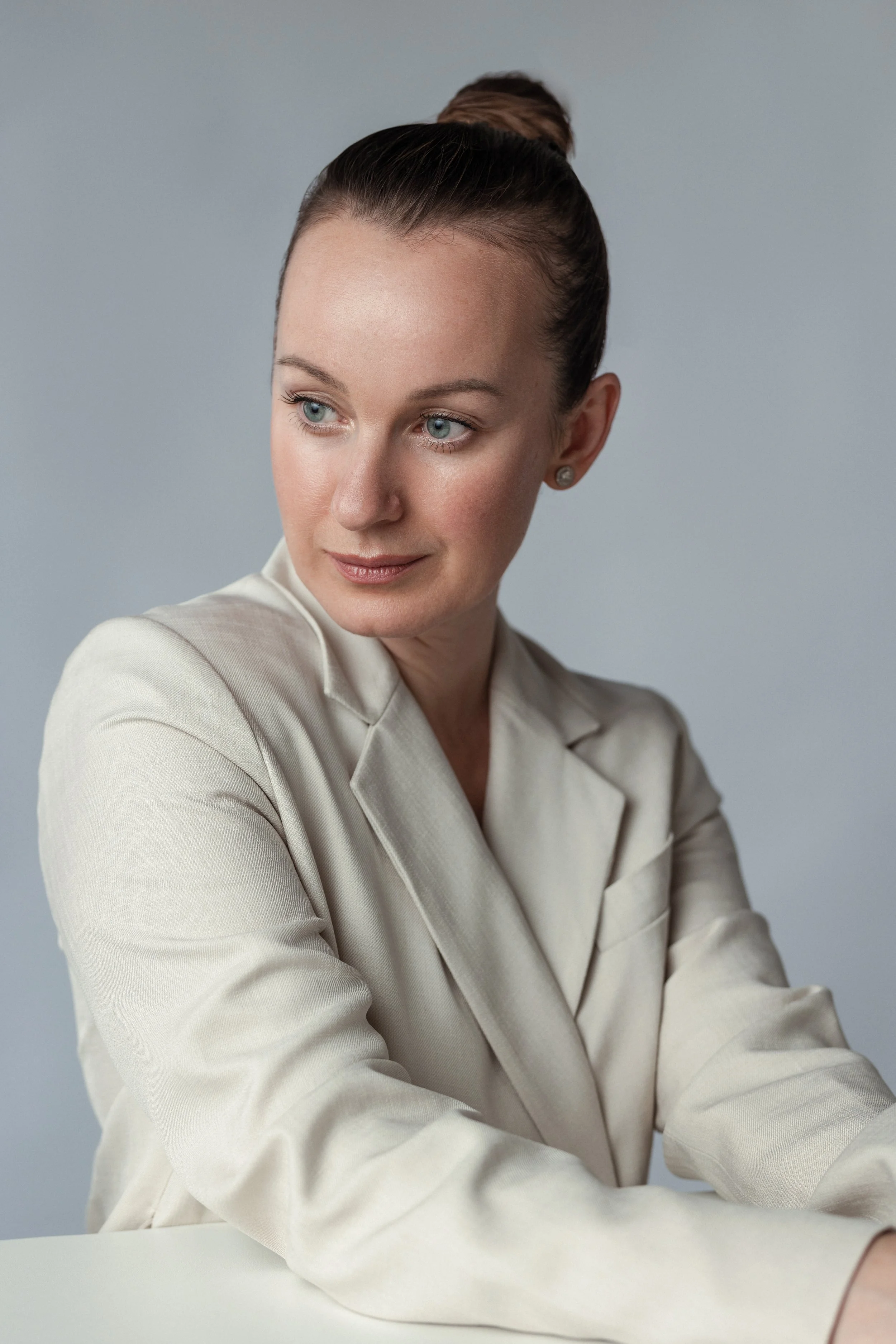 A professional corporate headshot: A woman with dark hair styled in a bun, wearing a beige blazer, seated with her arms crossed on a white table, against a plain light gray background.
