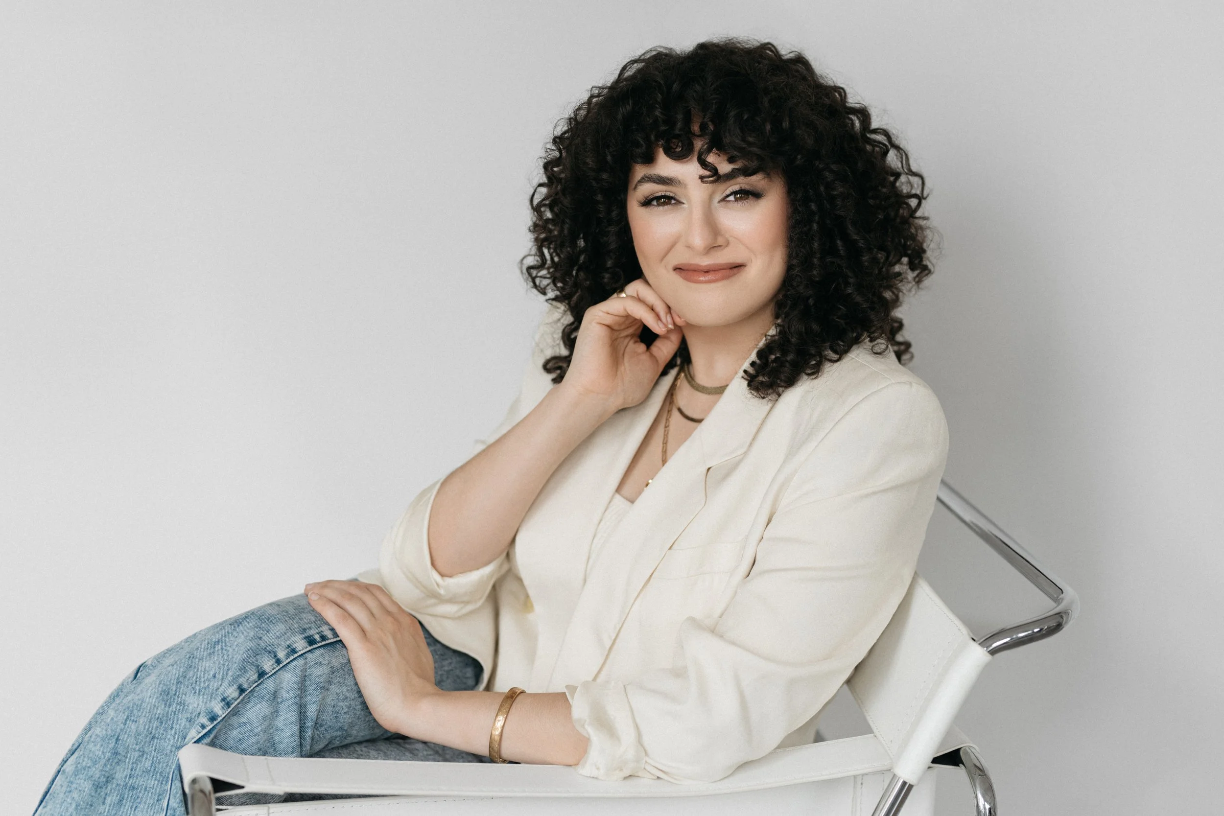 A professional corporate headshot of a woman with curly dark hair and light skin sitting on a white chair, wearing a cream-colored blazer, a gold bracelet, layered jewelry, and blue jeans, smiling softly and resting her chin on her hand.