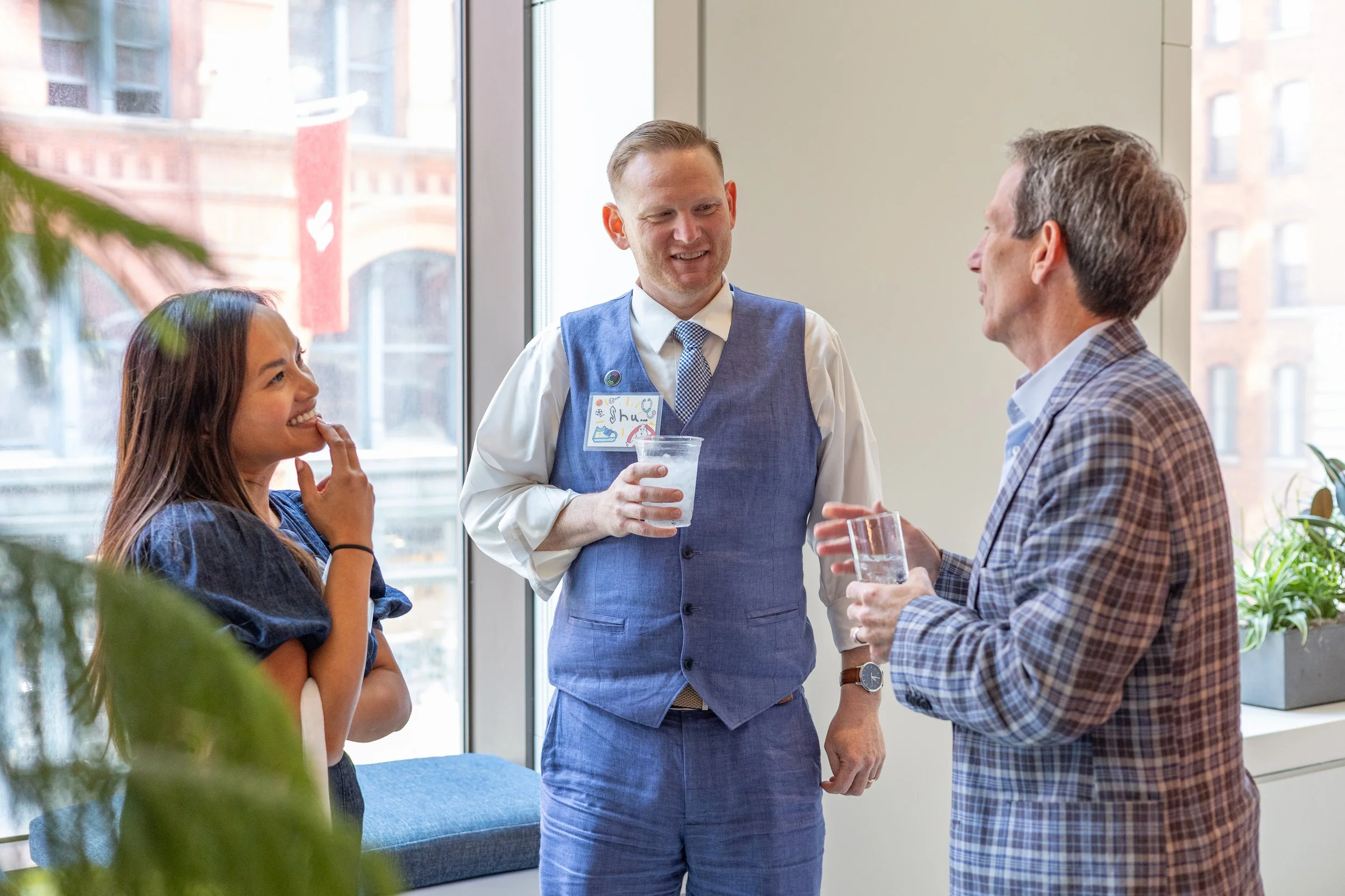A corporate event professional photo of three people chatting inside a building near a large window with an urban view; two are men, and one is a woman, all smiling and holding drinks. The man in the center is wearing a blue vest with a name tag.
