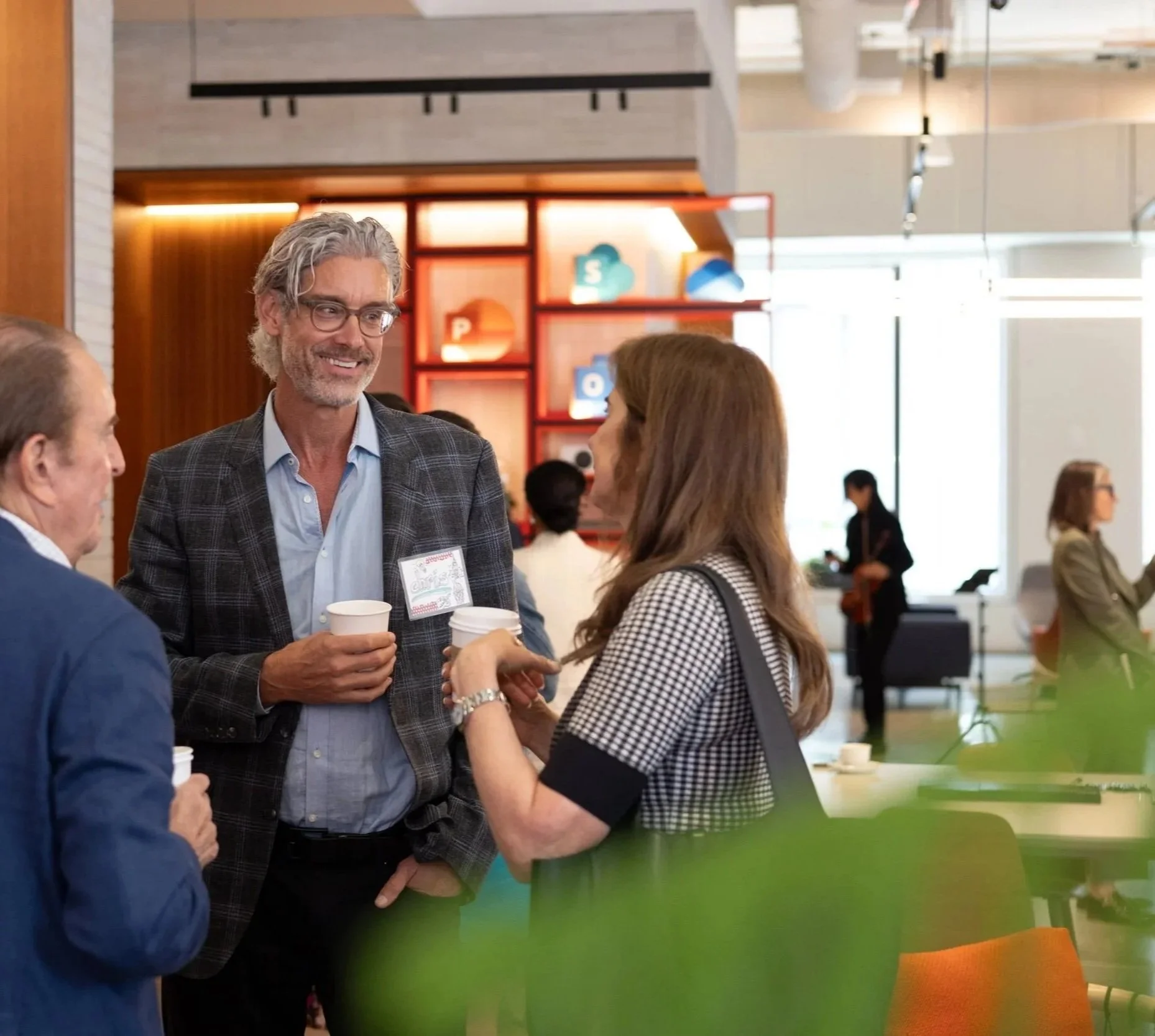 A corporate event photo of three people engaged in conversation at a professional networking event or conference in a modern, open space. In the background, more attendees are visible, and the environment features colorful decor and large windows.