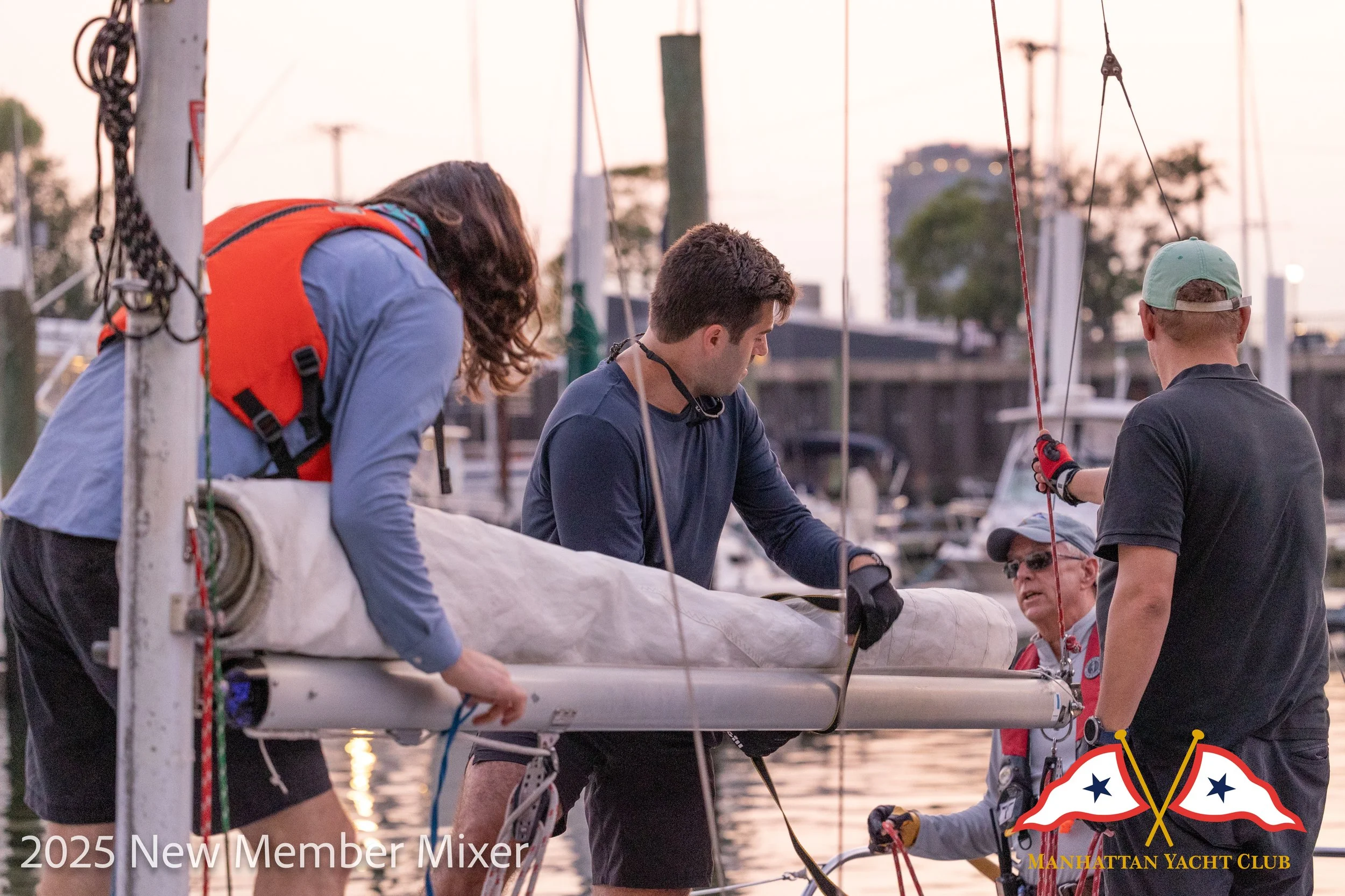A corporate event professional photo of a group of people preparing a sailboat at a marina during sunset.  There are boats and masts in the background. The bottom right corner has a logo: 'MANHATTAN YACHT CLUB'.