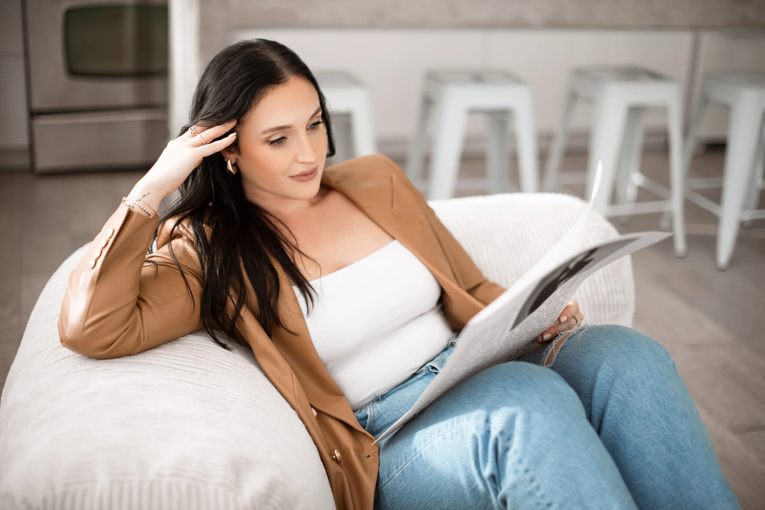A professional corporate headshot of a woman with dark hair sitting on a couch, reading a newspaper with her left hand resting on her forehead.
