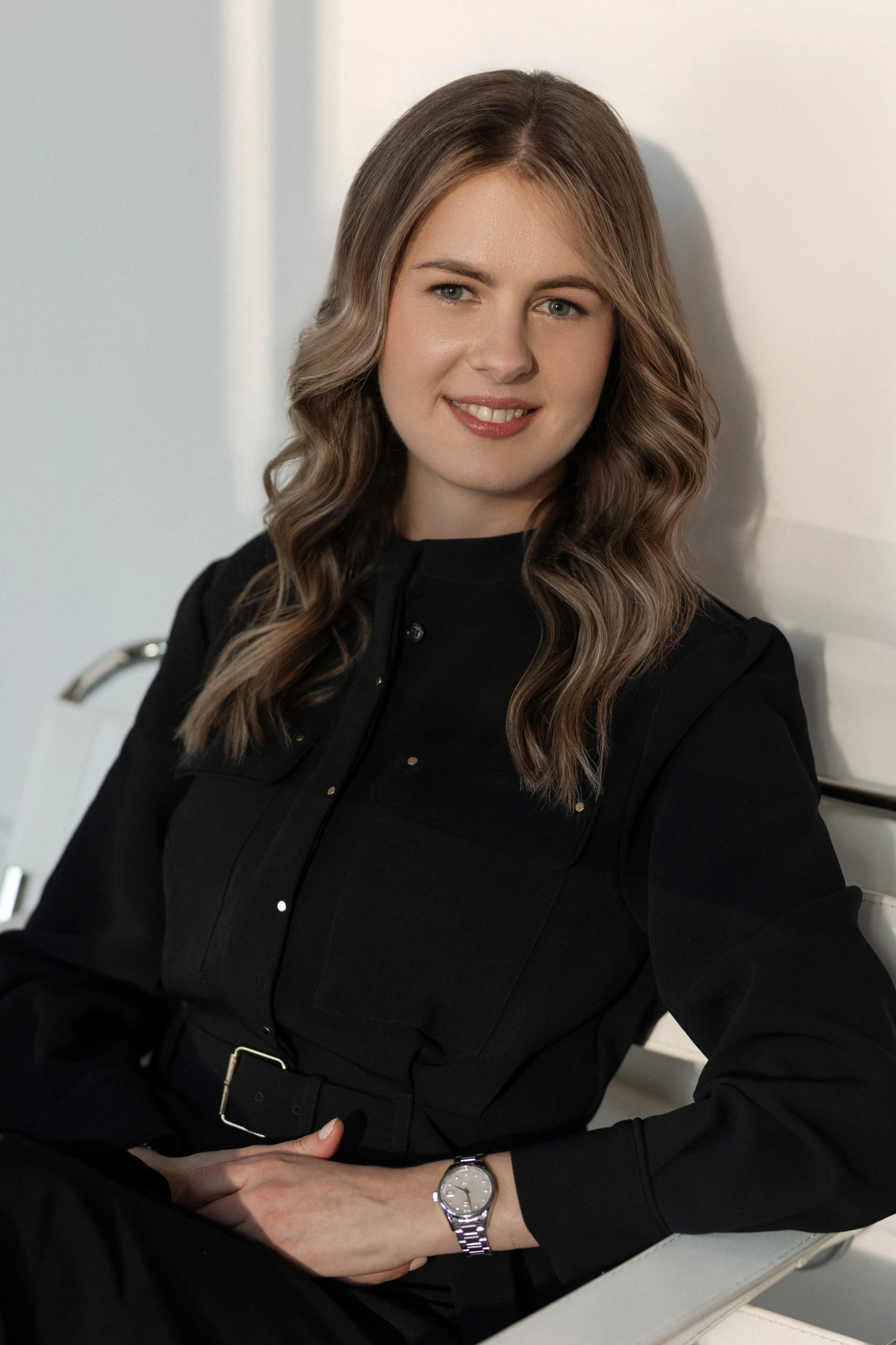 A professional corporate headshot of a woman with wavy brown hair and fair skin sitting at a desk, smiling, wearing a black top and a silver wristwatch.