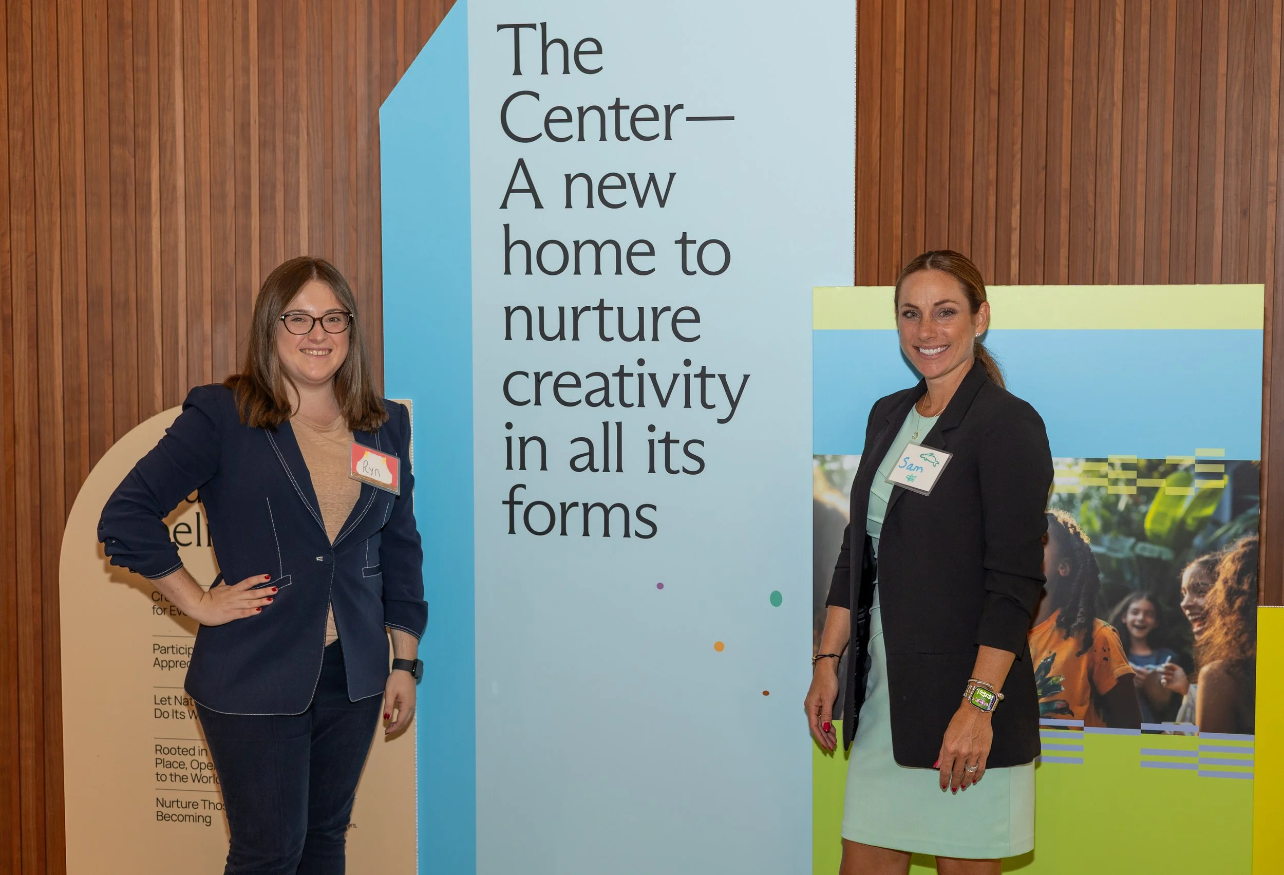 A corporate event professional photo of two women standing in front of a large sign that reads 'The Center—A new home to nurture creativity in all its forms'. They are smiling and wearing name tags, with a wooden wall background.