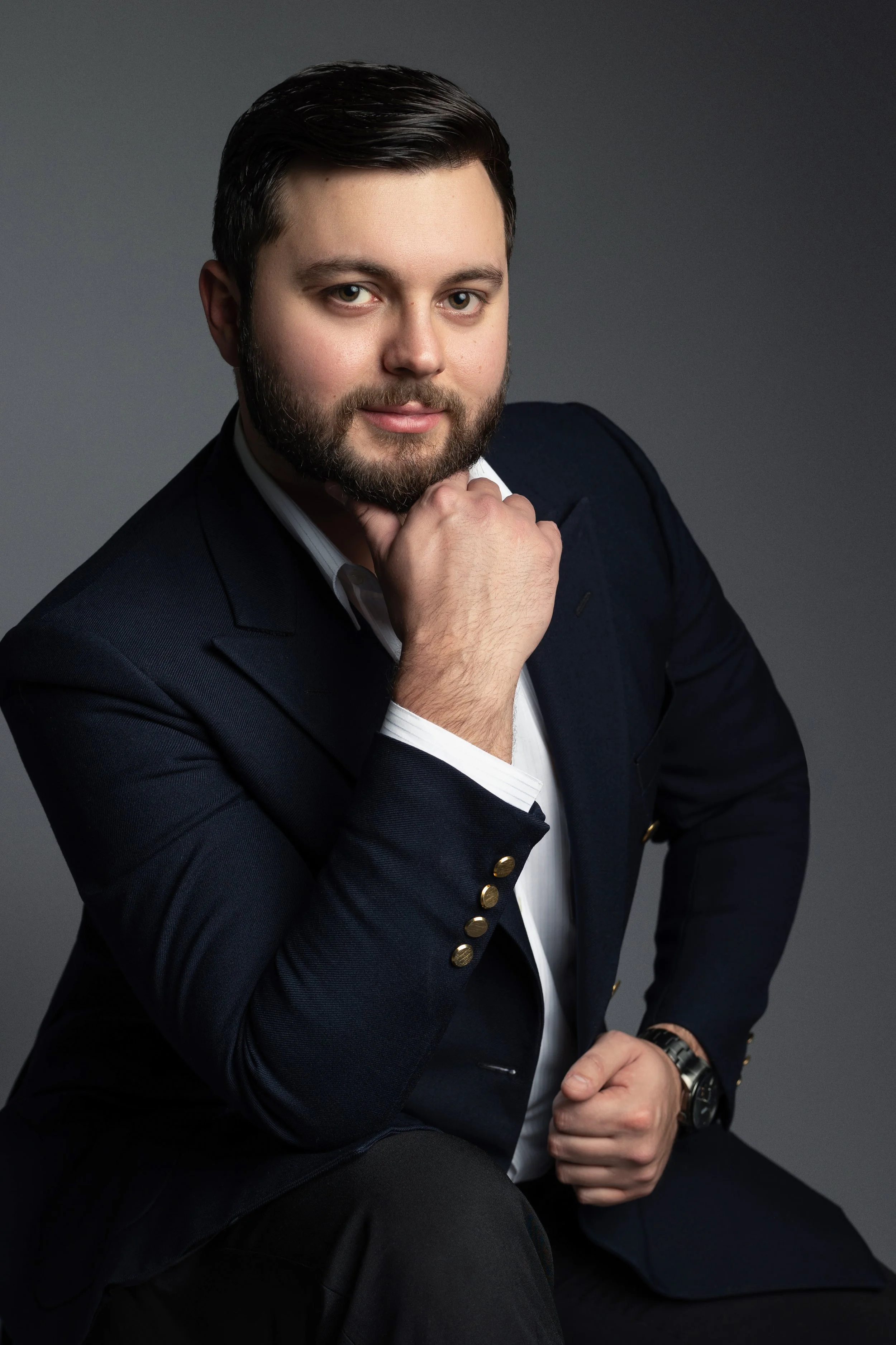 A portrait of a man with dark hair and a beard, wearing a dark blazer with gold buttons over a white shirt, sitting against a gray background. He is resting his chin on his right hand and looking at the camera.
