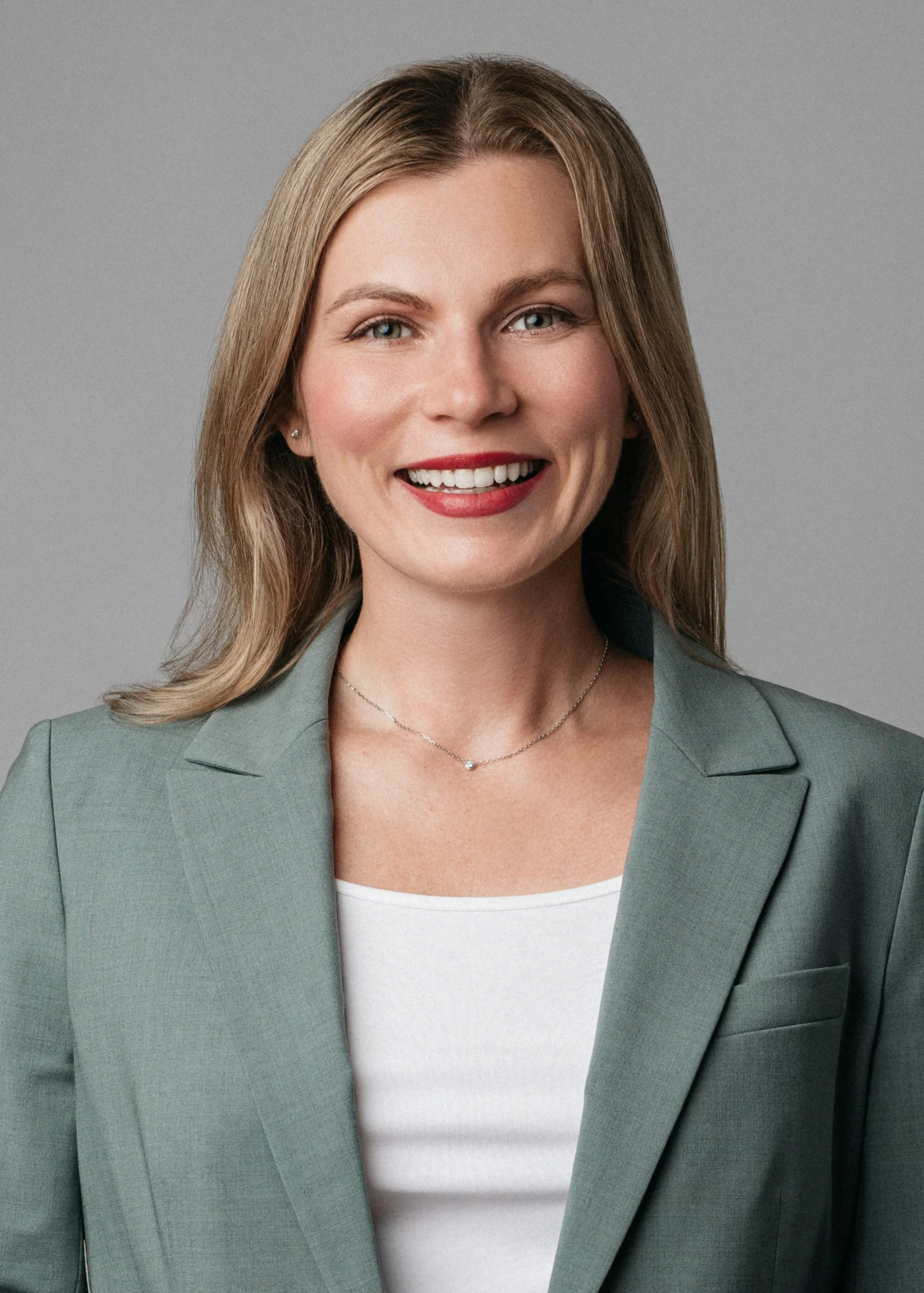A professional corporate headshot of a professional woman with blonde hair smiling, wearing a green blazer, white top, small earrings, and a delicate necklace, against a gray background.