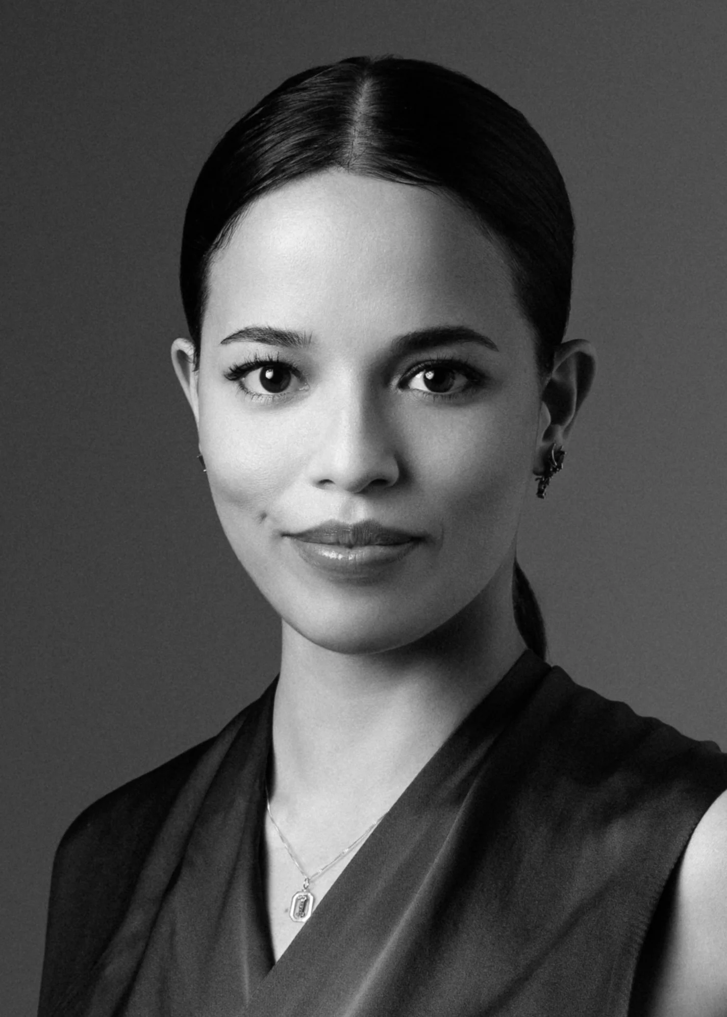A professional corporate headshot of a black-and-white portrait of a woman with dark, pulled-back hair, wearing earrings and a necklace, smiling slightly, dressed in dark clothing, standing against a plain background.