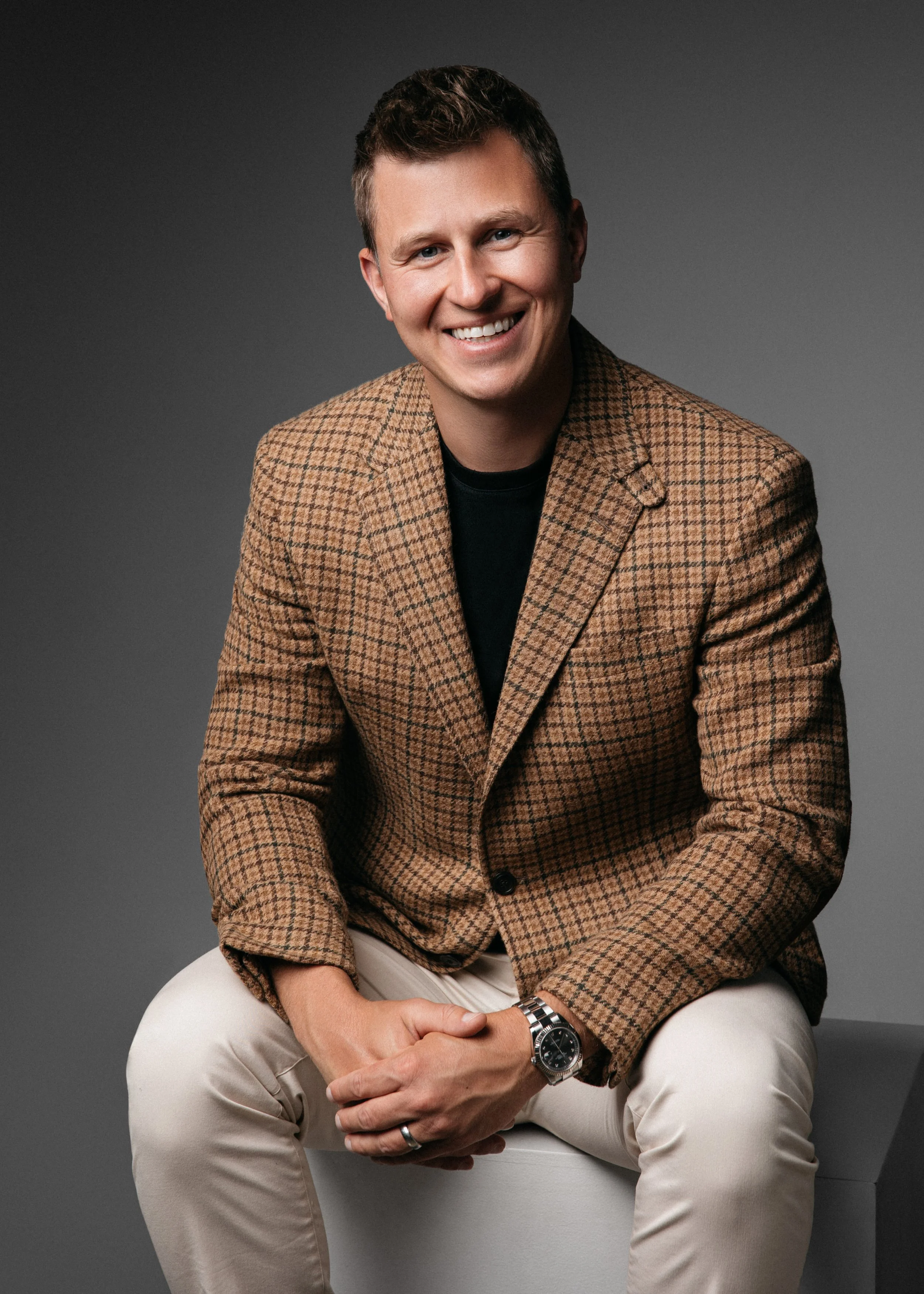 A professional corporate headshot of a smiling man wearing a brown checkered blazer, black shirt, and beige pants, sitting on a white bench against a gray background.