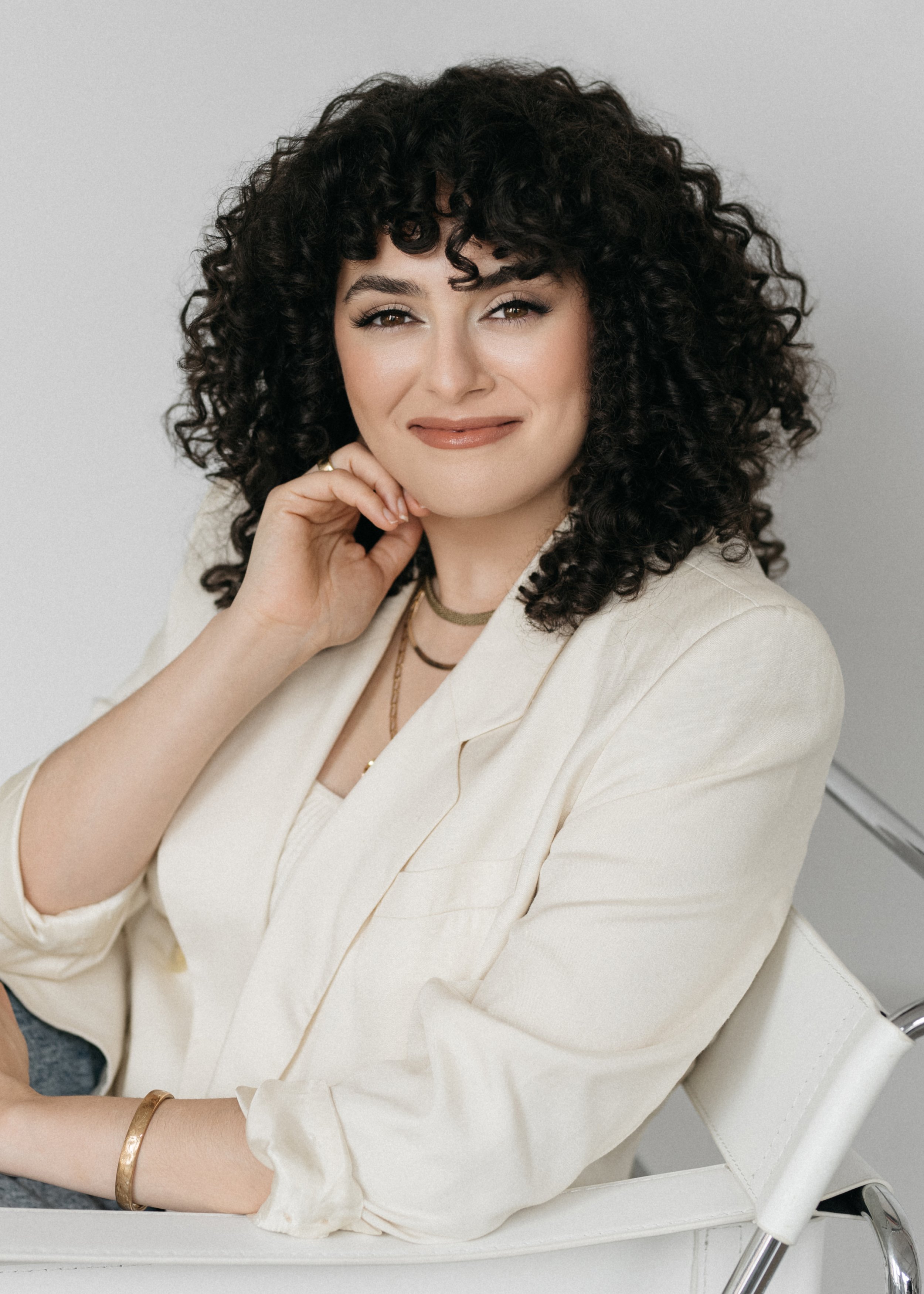 A professional corporate headshot of a young woman with dark curly hair and light makeup, wearing a cream blazer and gold jewelry, sitting on a white chair against a plain gray background.