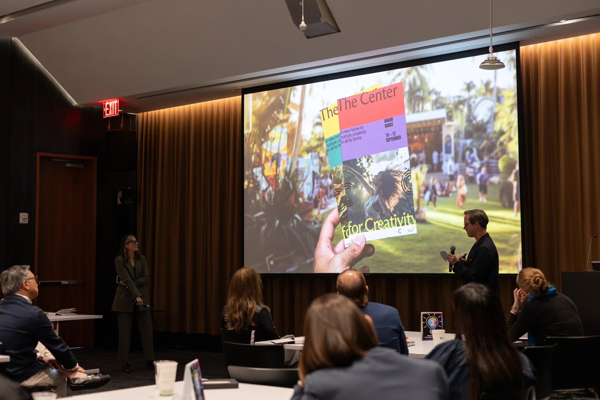 A corporate event: a conference room with a large screen displaying a colorful flyer for an event. People are seated and standing, observing two presenters at the front of the room, one holding a microphone and the other standing near the screen.