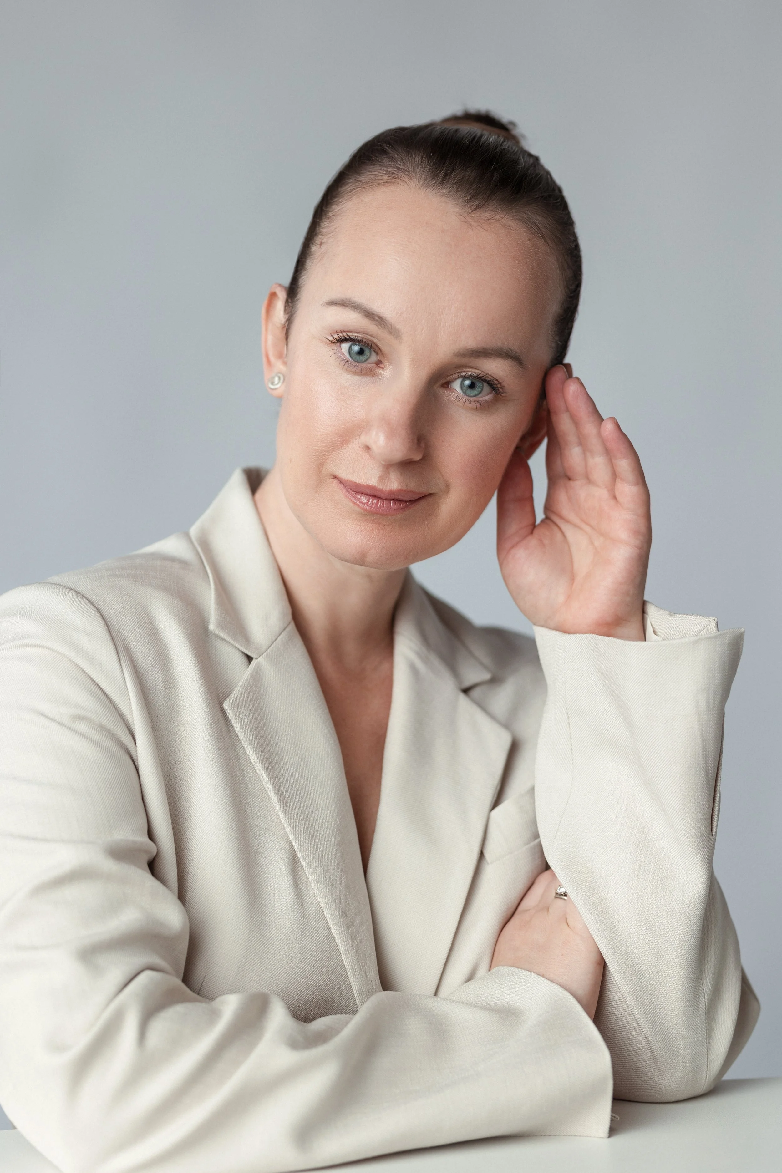 A professional corporate headshot of a woman with blue eyes and dark hair styled in a high bun, wearing a beige blazer and pearl earrings, smiling softly with her hand resting on her face, against a plain light background.