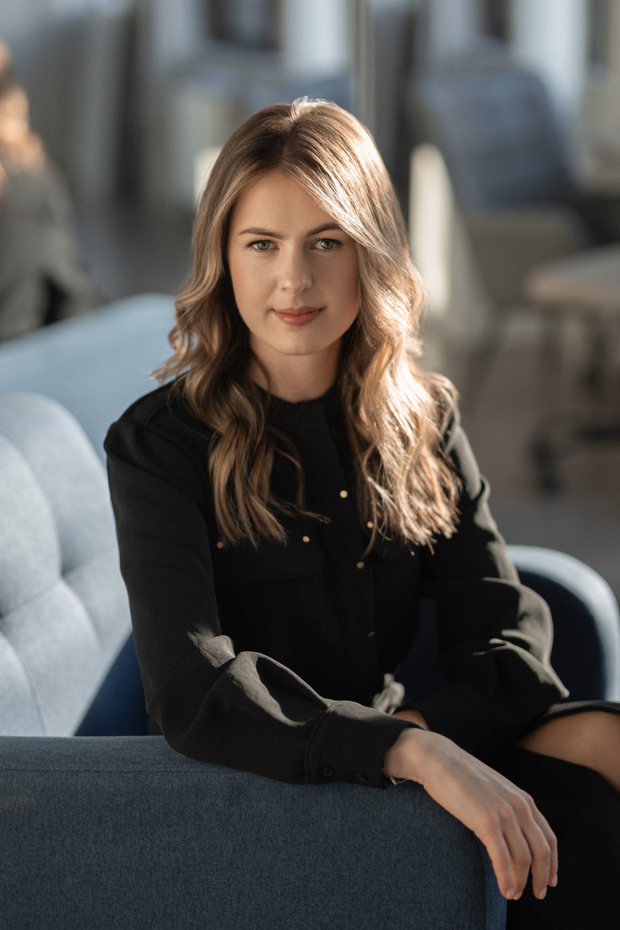 A professional corporate headshot of a young woman with wavy brown hair, wearing a black top, sitting on a blue couch with a blurred background of a modern interior.