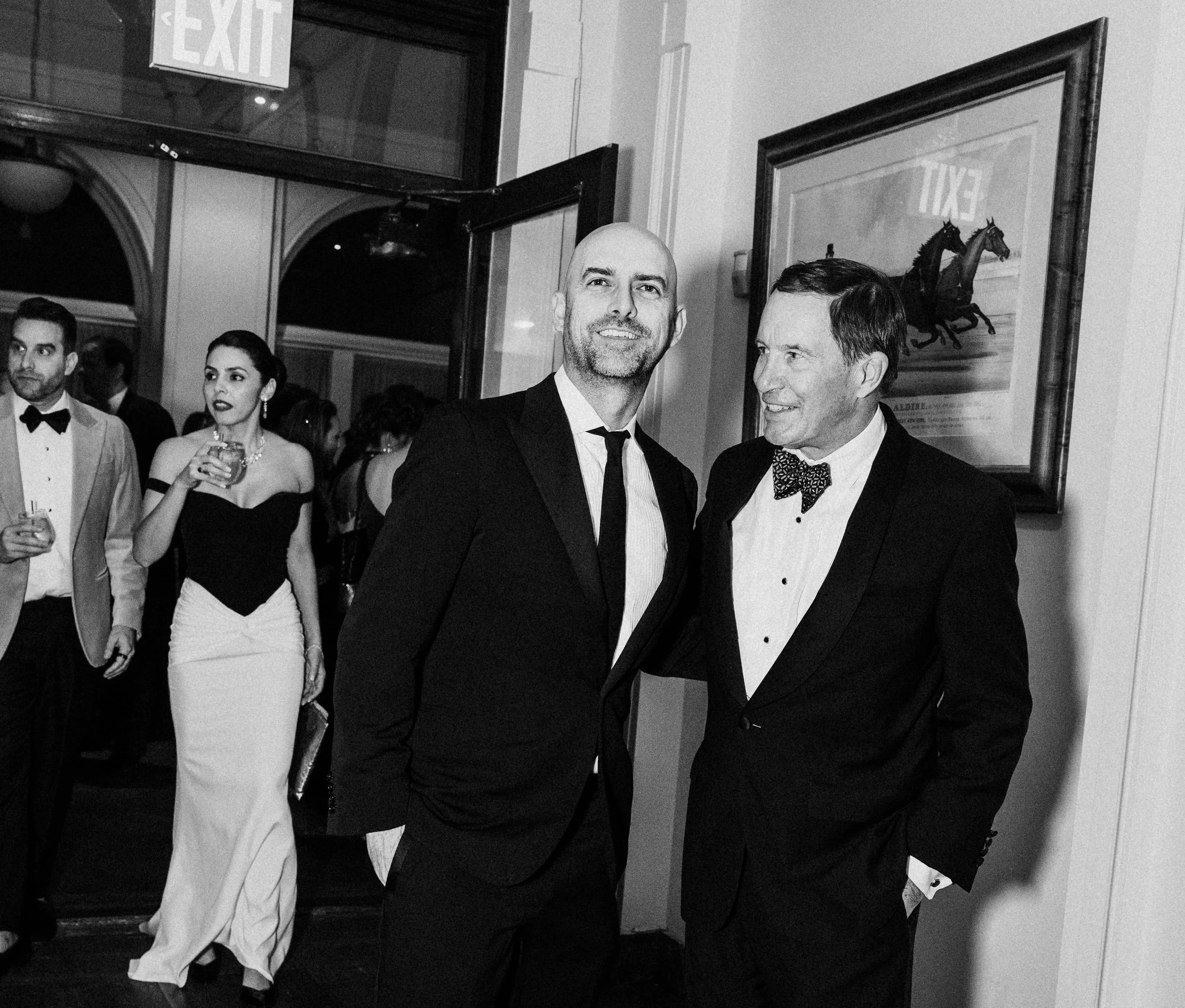 A corporate event professional photo of a black-and-white photo of a formal party with men in tuxedos and women in elegant dresses indoors. Two men are in the foreground, smiling and talking.