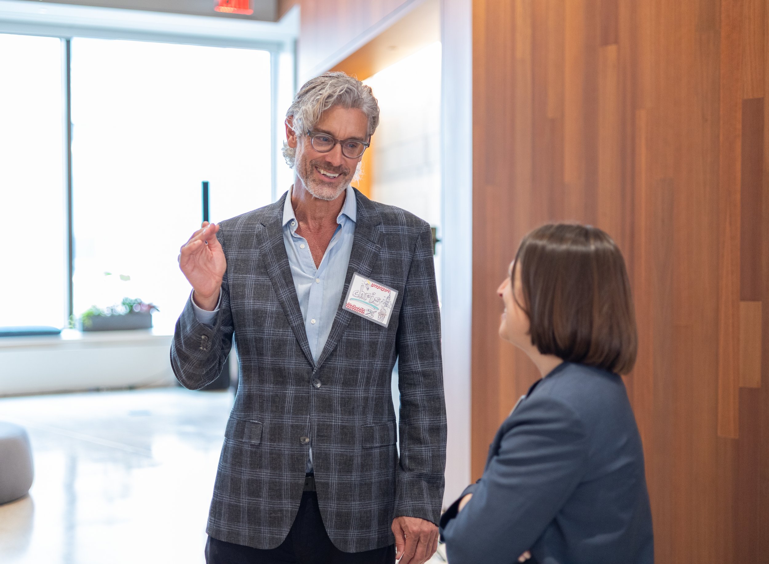 A corporate event professional photo of a man with glasses and gray hair wearing a gray plaid blazer and light blue shirt, smiling and talking with a woman with brown hair in a navy blazer in an indoor setting with wooden walls and large windows.