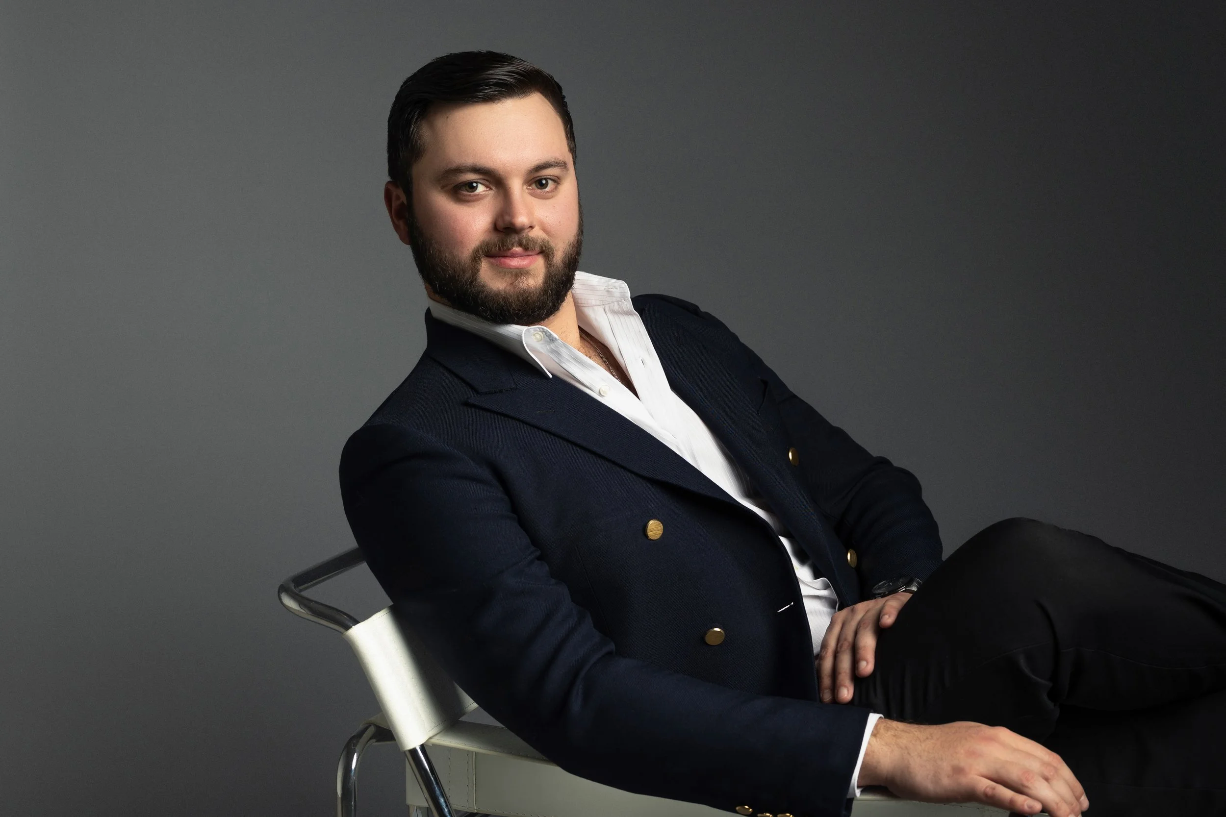 A professional corporate headshot of a man with dark hair and a beard, wearing a navy blazer and white shirt, sitting on a white chair against a plain gray background.