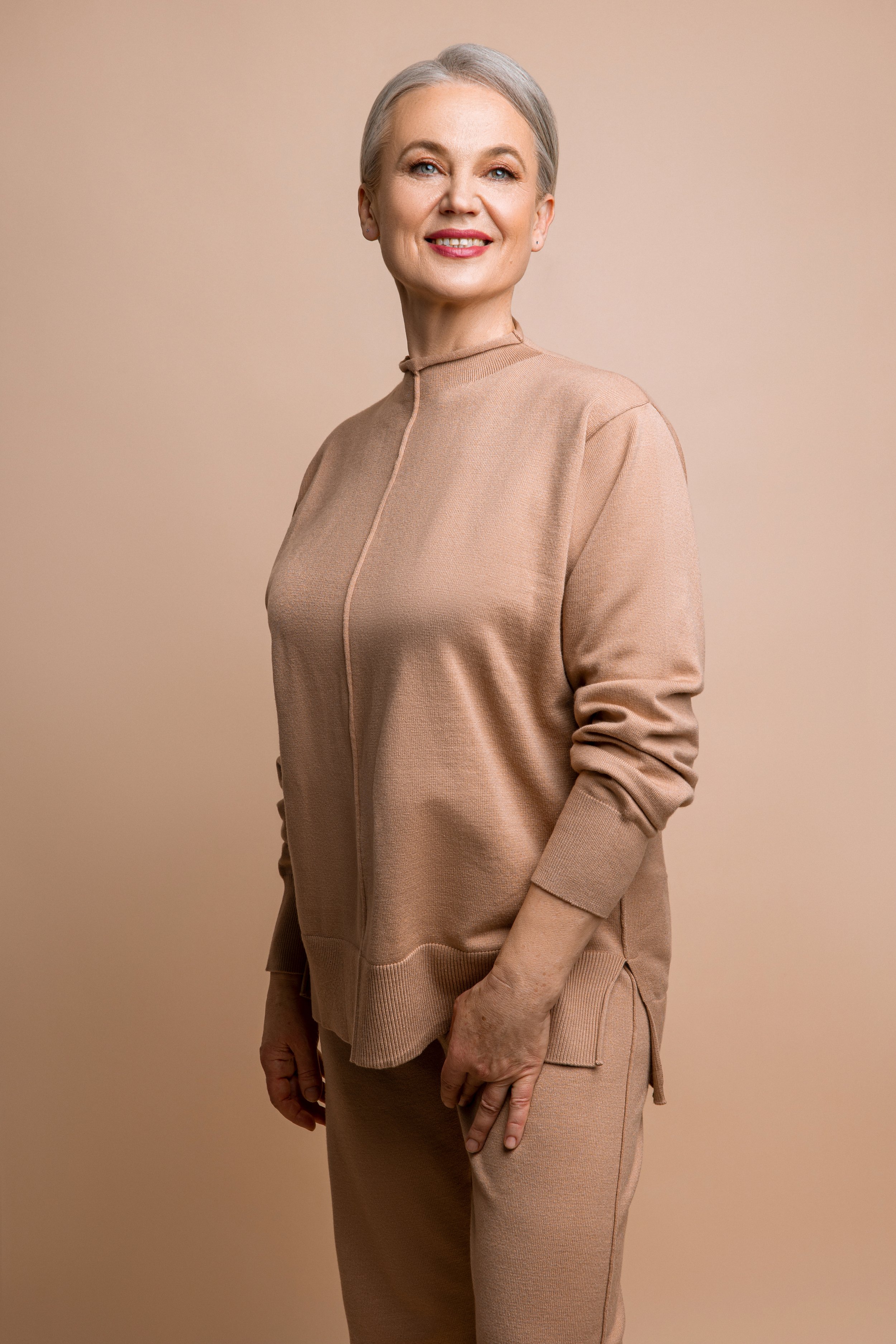 A professional corporate headshot of an elderly woman with short gray hair, smiling, wearing a beige zip-up top and beige pants, standing against a plain beige background.